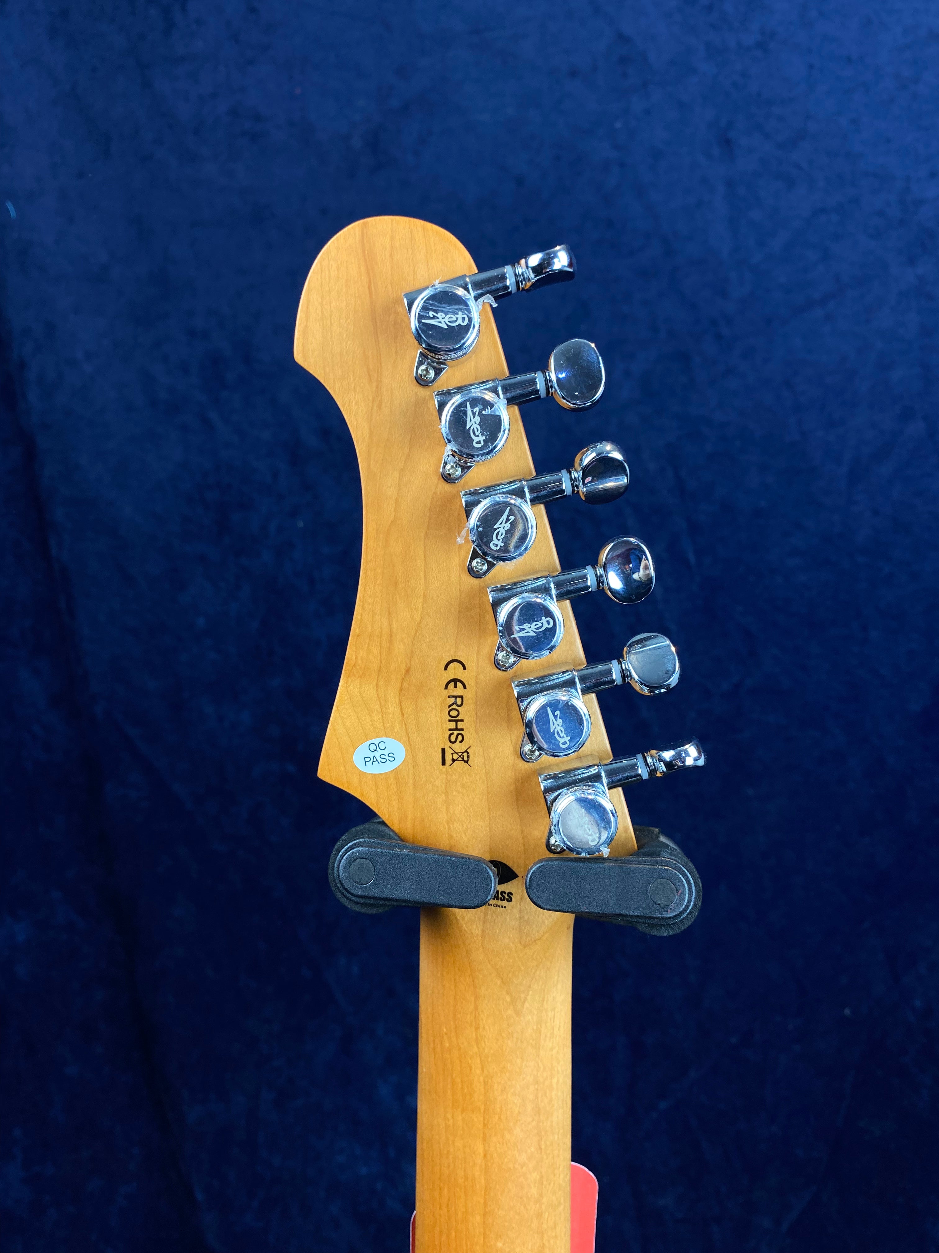 Six chrome tuning pegs line the light wood headstock of an electric guitar, with QC PASS and certification labels visible on the back.