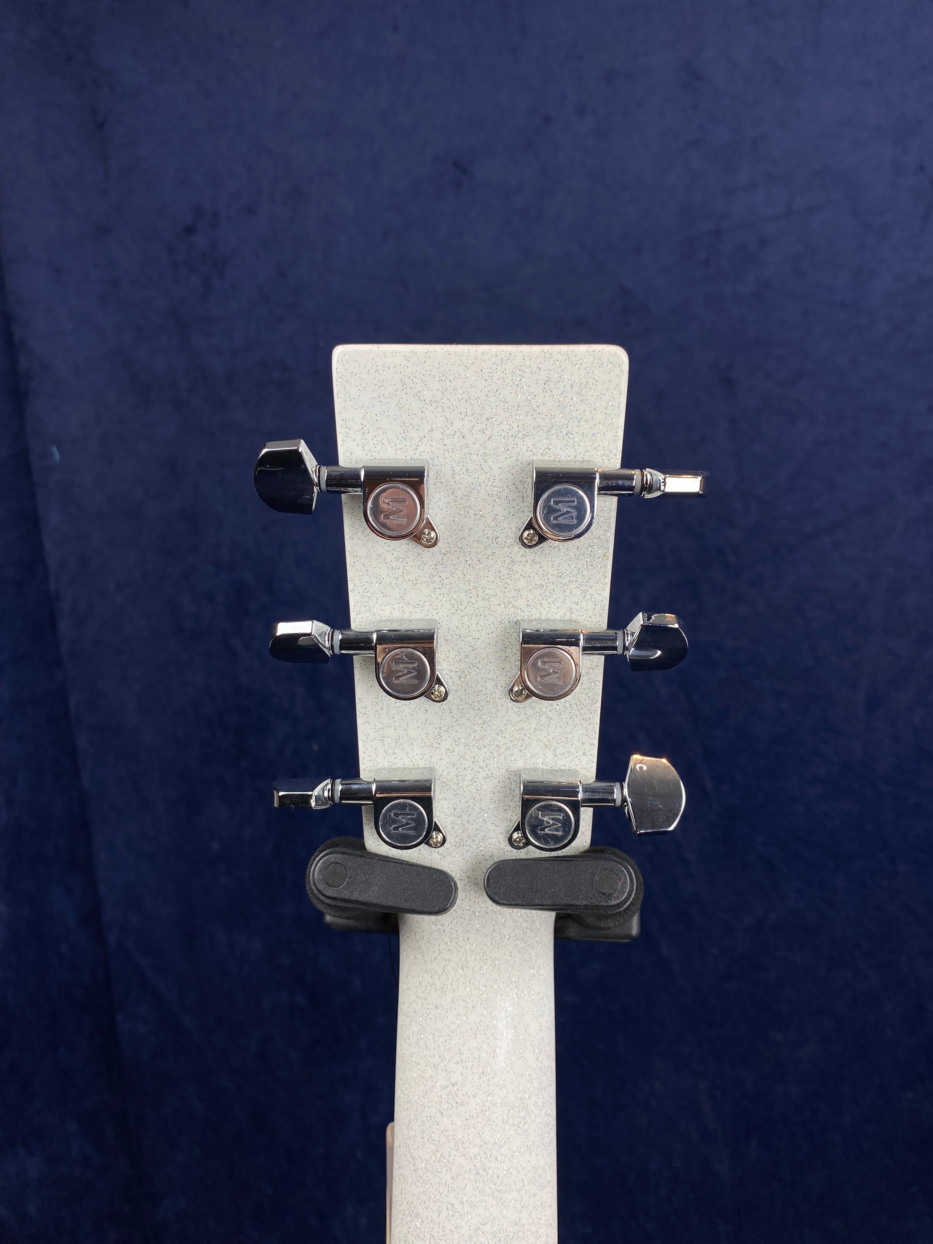 Six chrome tuning pegs on the headstock of a white acoustic guitar, viewed from the back against a dark blue background.