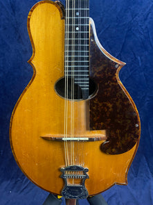 Carved wooden mandolin with a distinctive asymmetrical body shape, eight strings, a dark tortoiseshell-patterned pickguard, and a decorative metal tailpiece.