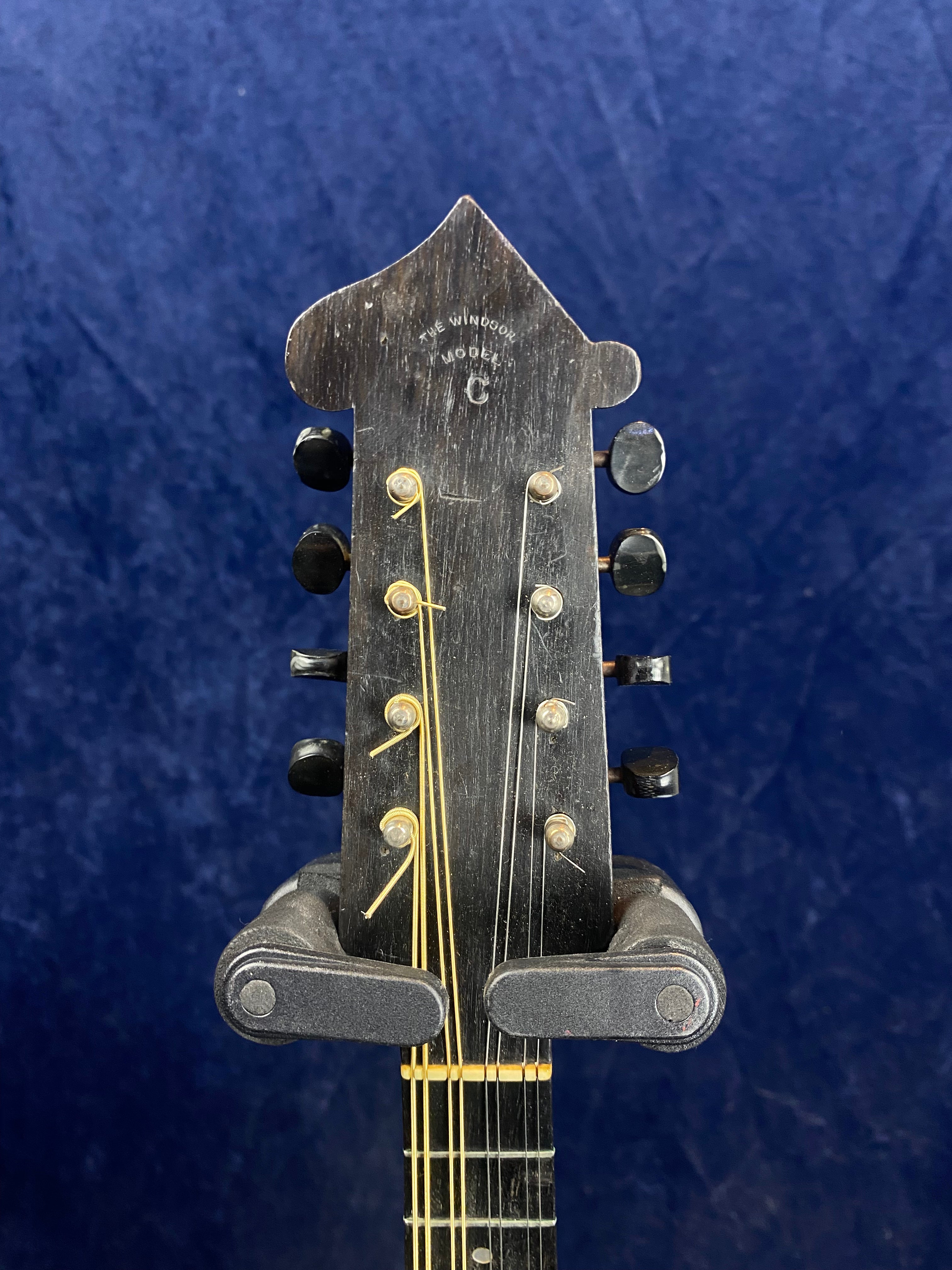 Black mandolin headstock with eight tuning pegs, gold strings, and “The Windsor Model C” text at the top, set against a dark blue background.