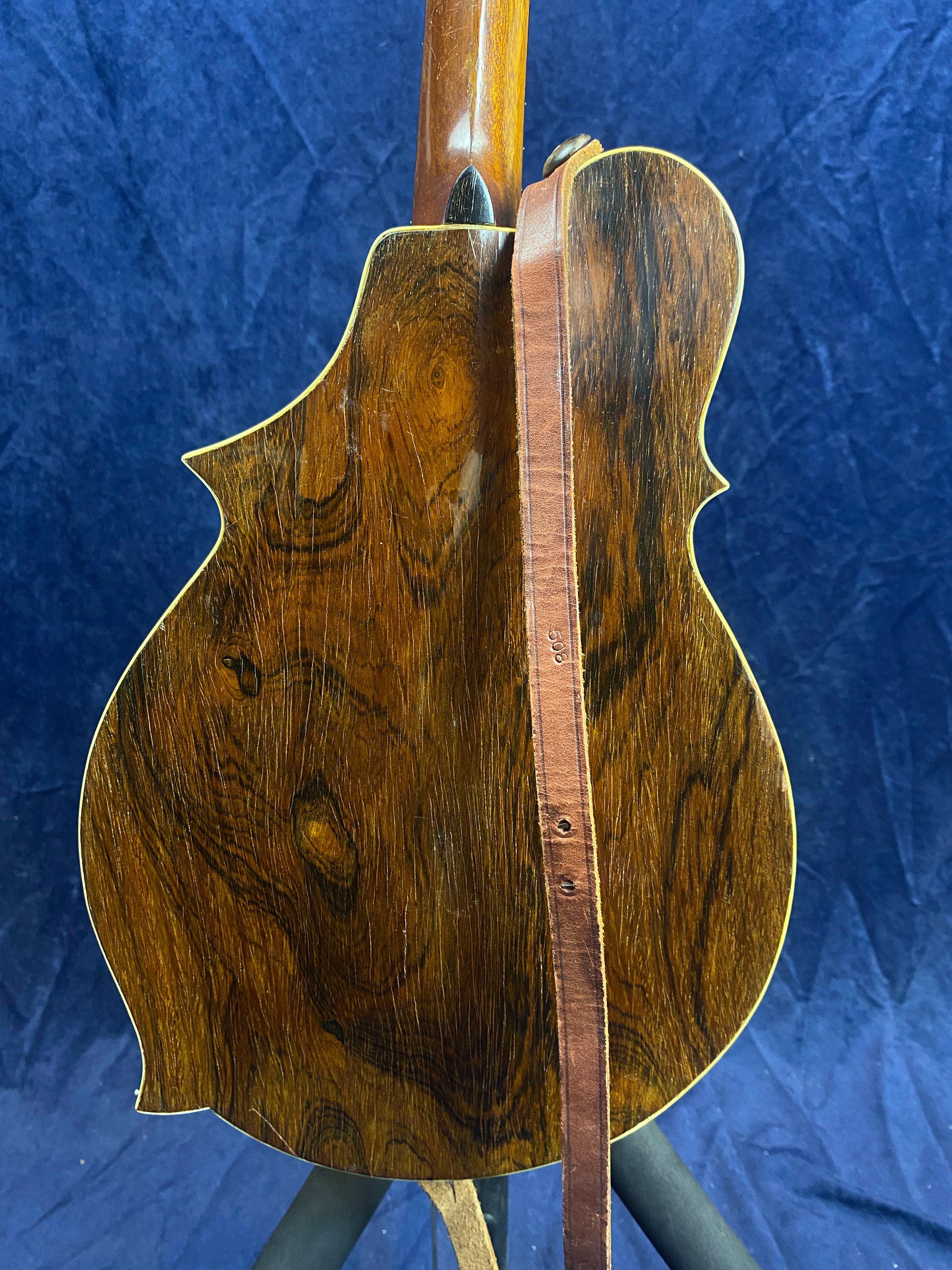 Mandolin with a glossy dark wood back, visible rich grain patterns, and a brown leather strap draped over it, set against a blue background.