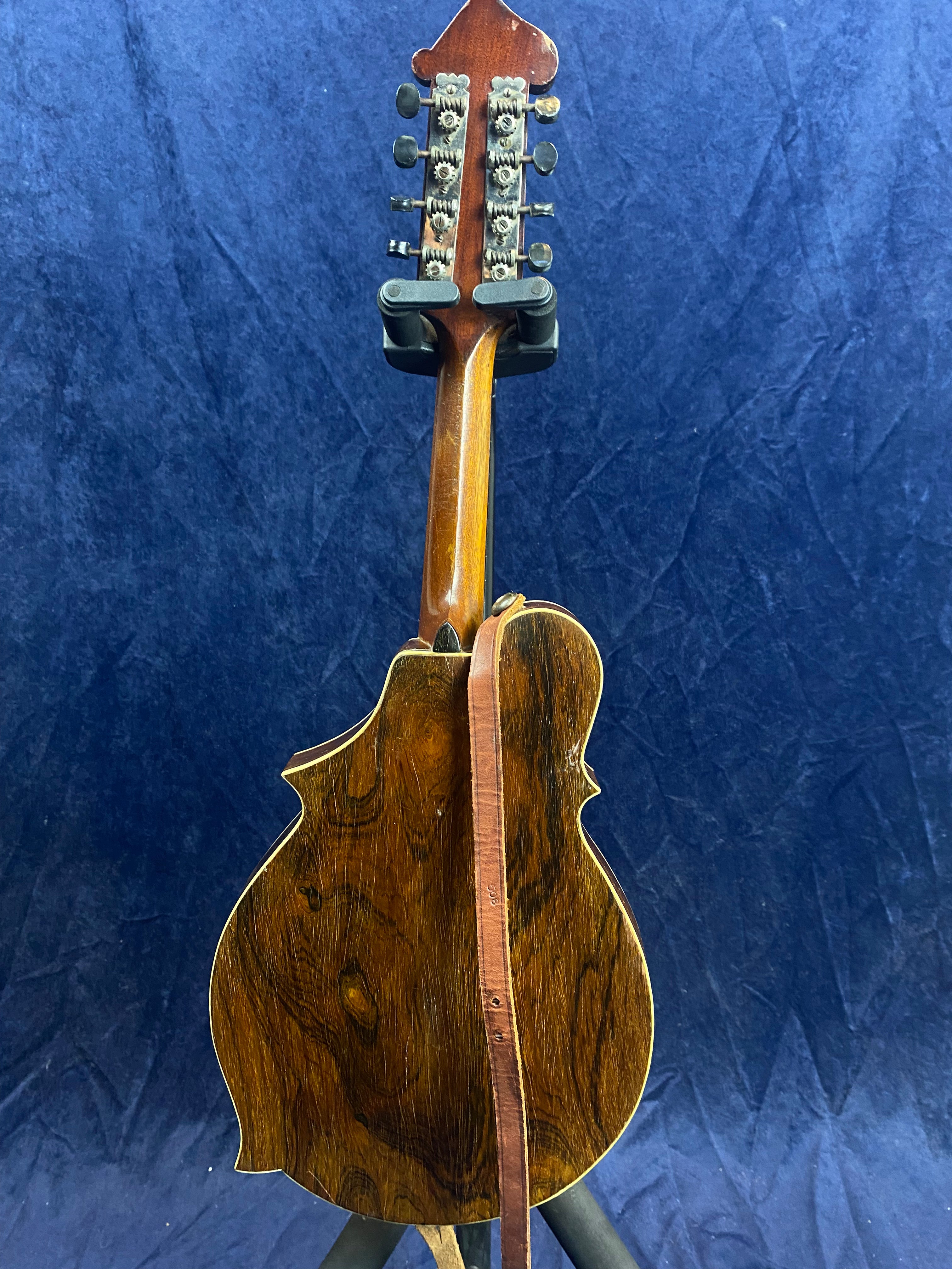 Dark wood mandolin shown from the back, displaying rich grain patterns, tuning pegs, and a brown leather strap hanging down the neck.