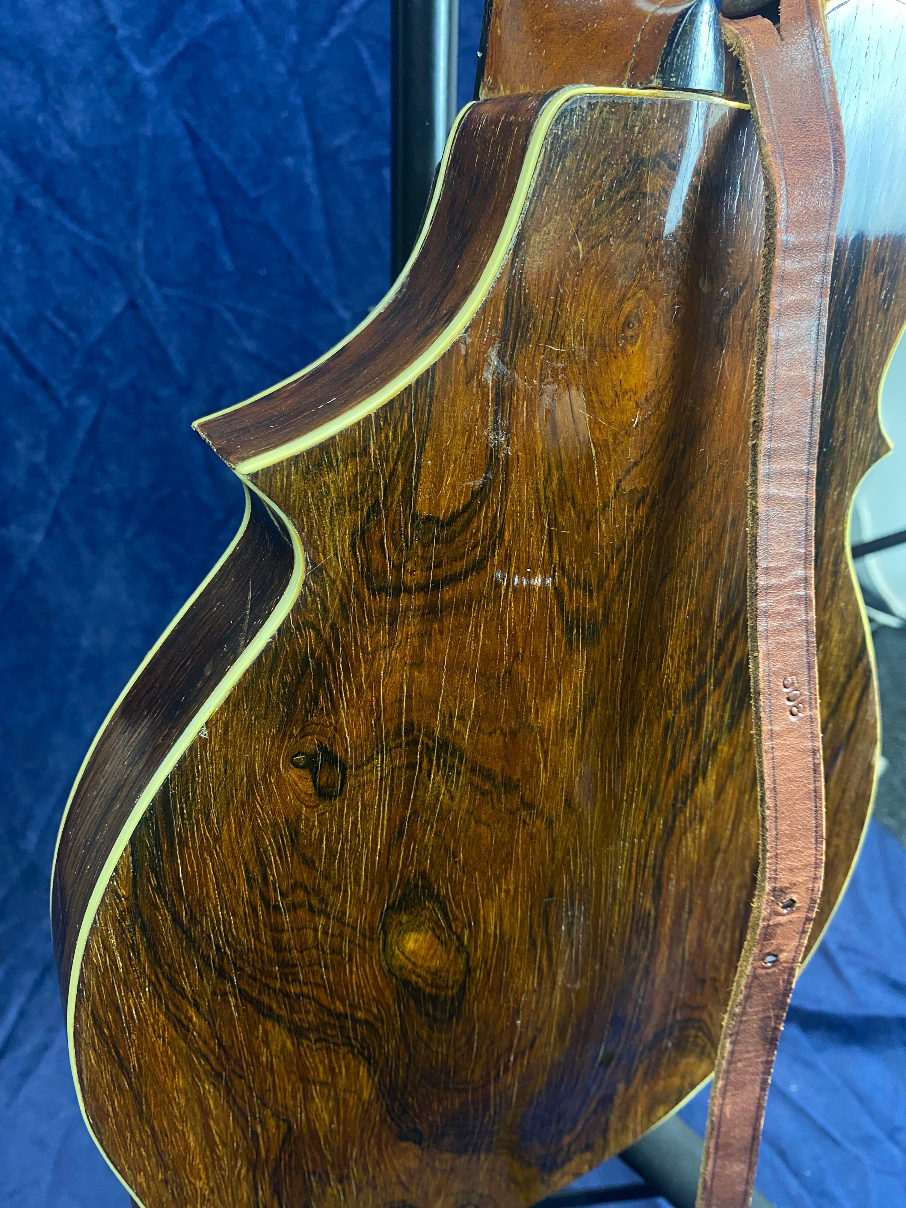 Curved back of a wooden mandolin with a glossy finish, natural wood grain, cream edge binding, and a brown leather strap attached.
