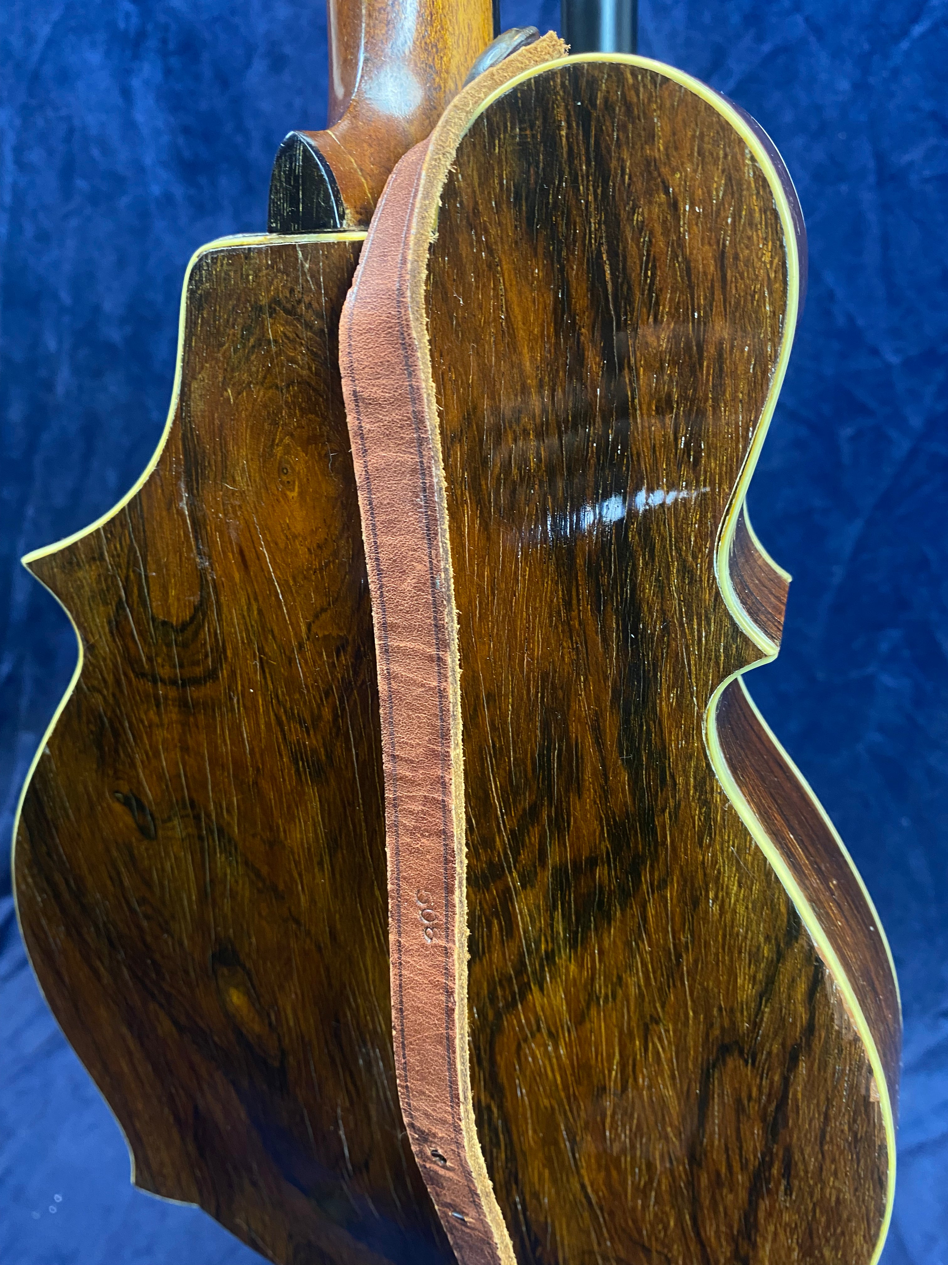 A brown wooden mandolin with a glossy finish and light yellow trim, shown from the back with a tan leather strap attached along the side.