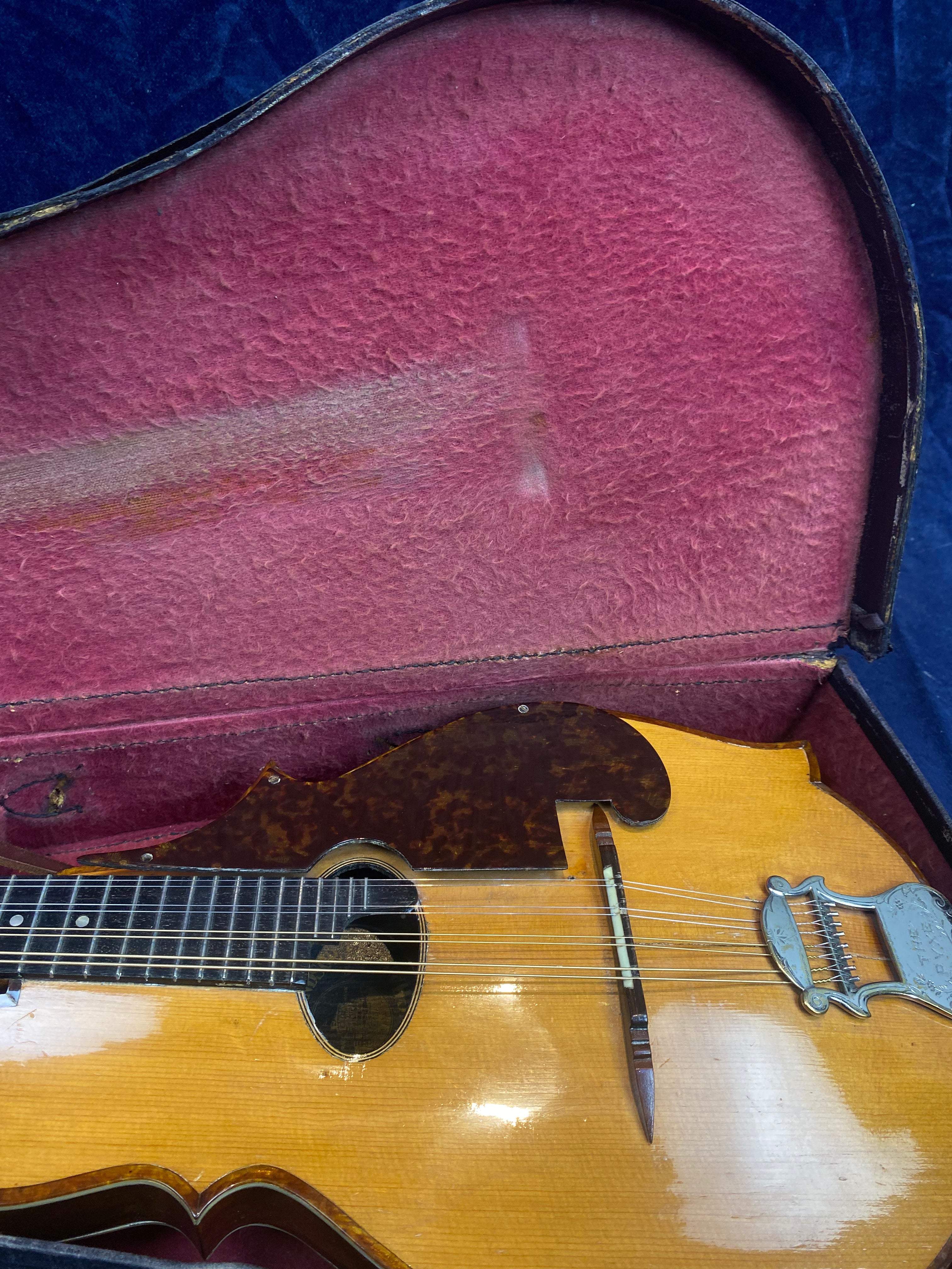 Vintage mandolin with a yellowed wooden body, tortoiseshell pickguard, and metal tailpiece, resting inside a plush red-lined hard case.