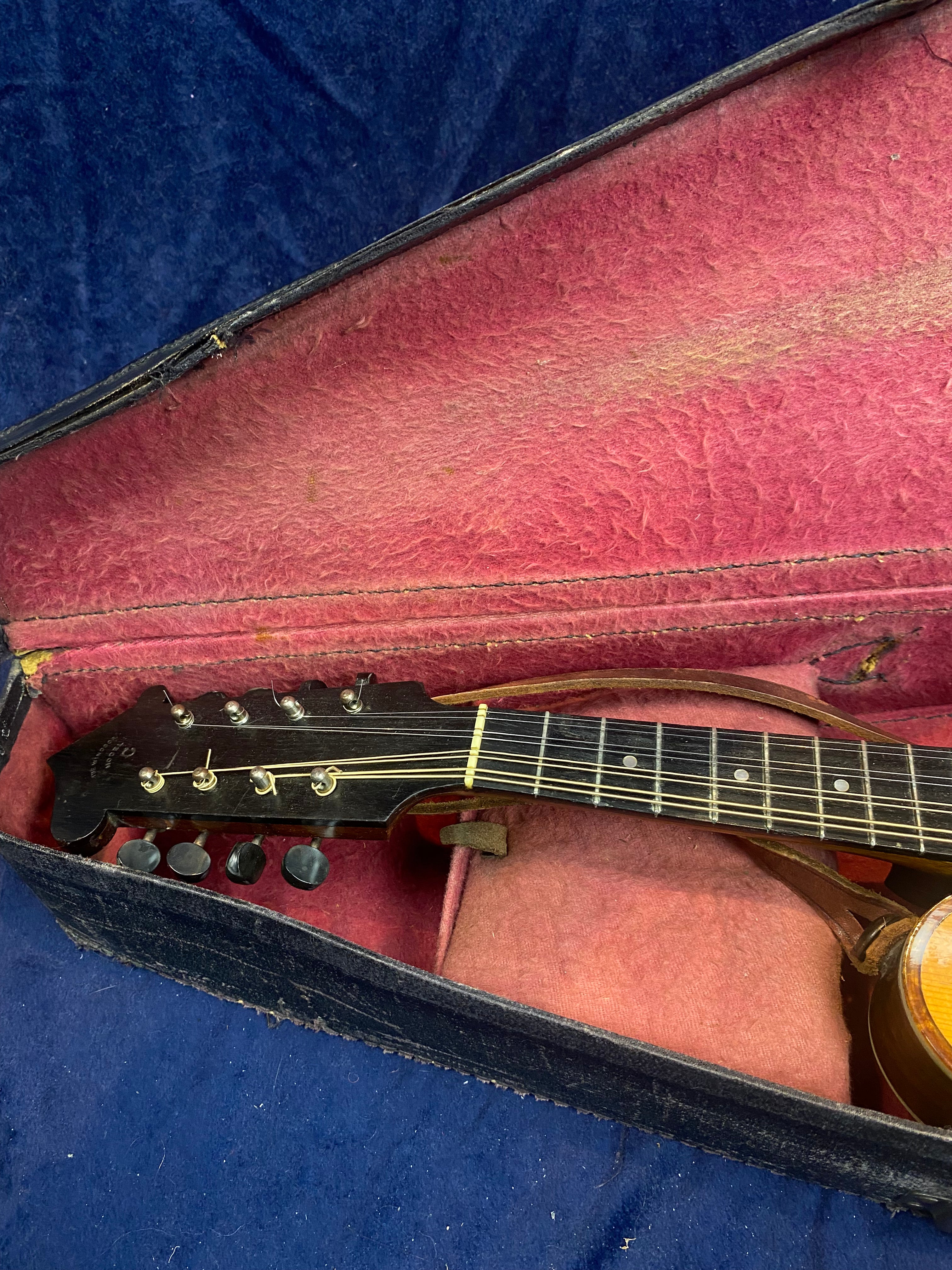 A mandolin rests in a worn, open case lined with faded pink fabric; the instrument's dark headstock and part of the neck are visible.