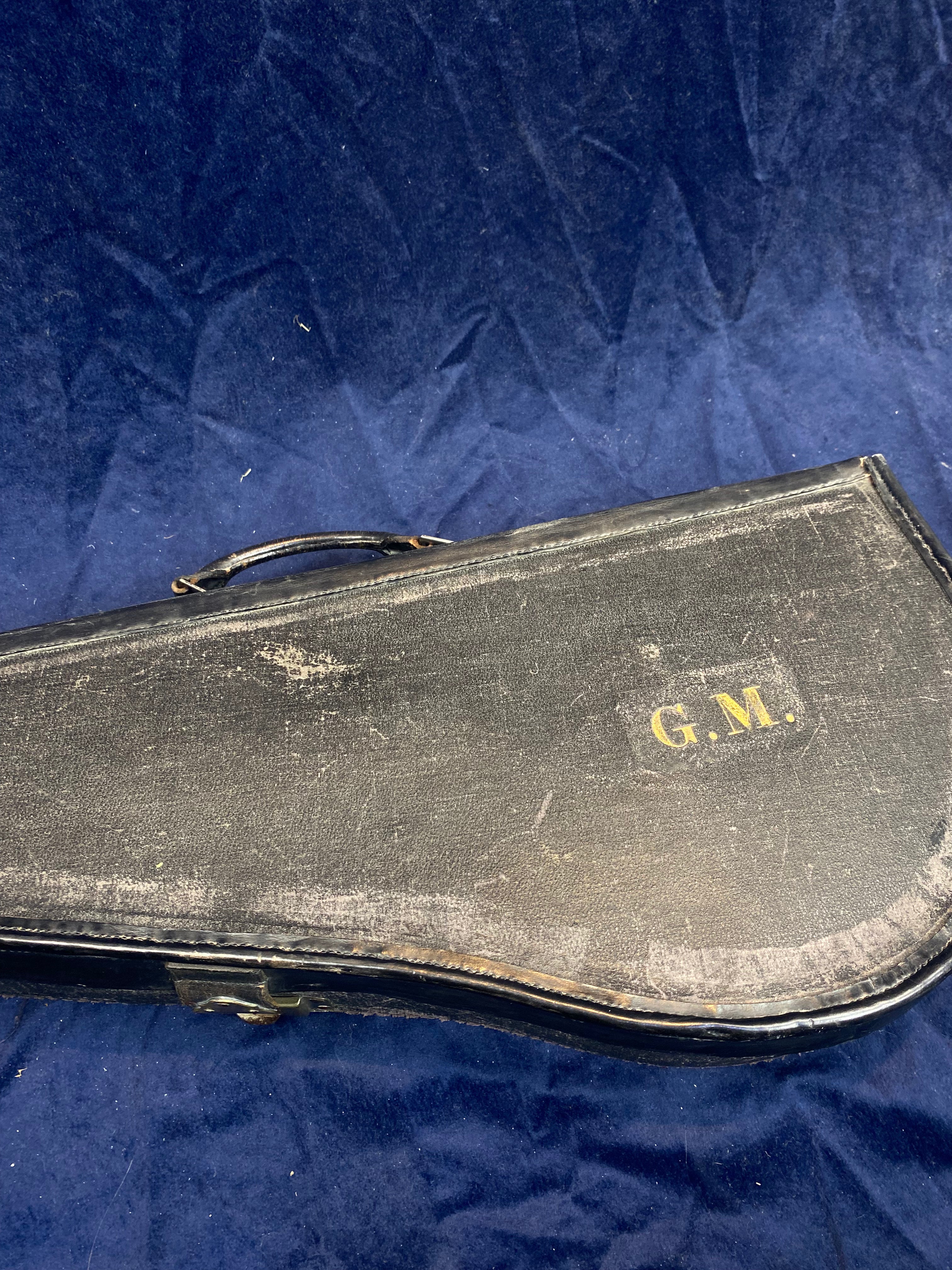 Worn black mandolin case with gold initials "G.M." on the lid, sitting on a dark blue textured surface.