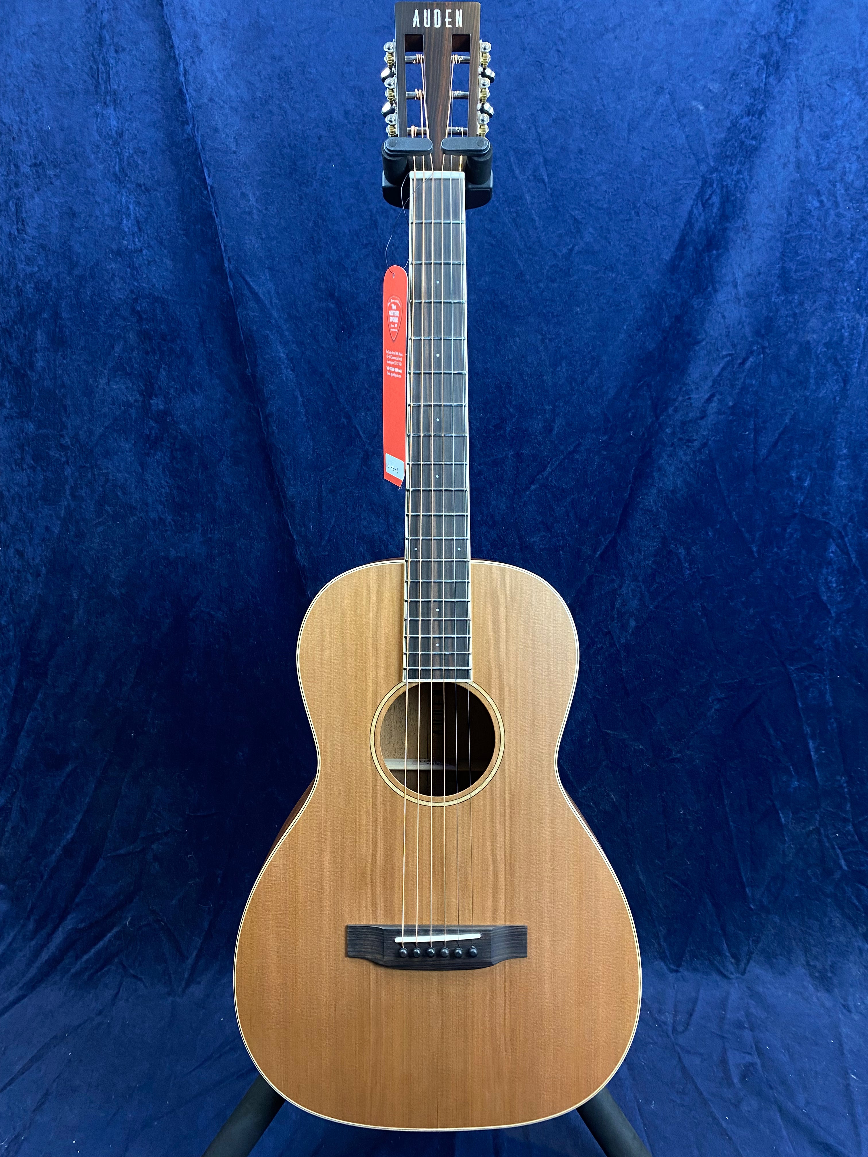 Auden acoustic guitar with a natural wood finish, six strings, dark fretboard, and a red tag hanging from the headstock against a blue background.