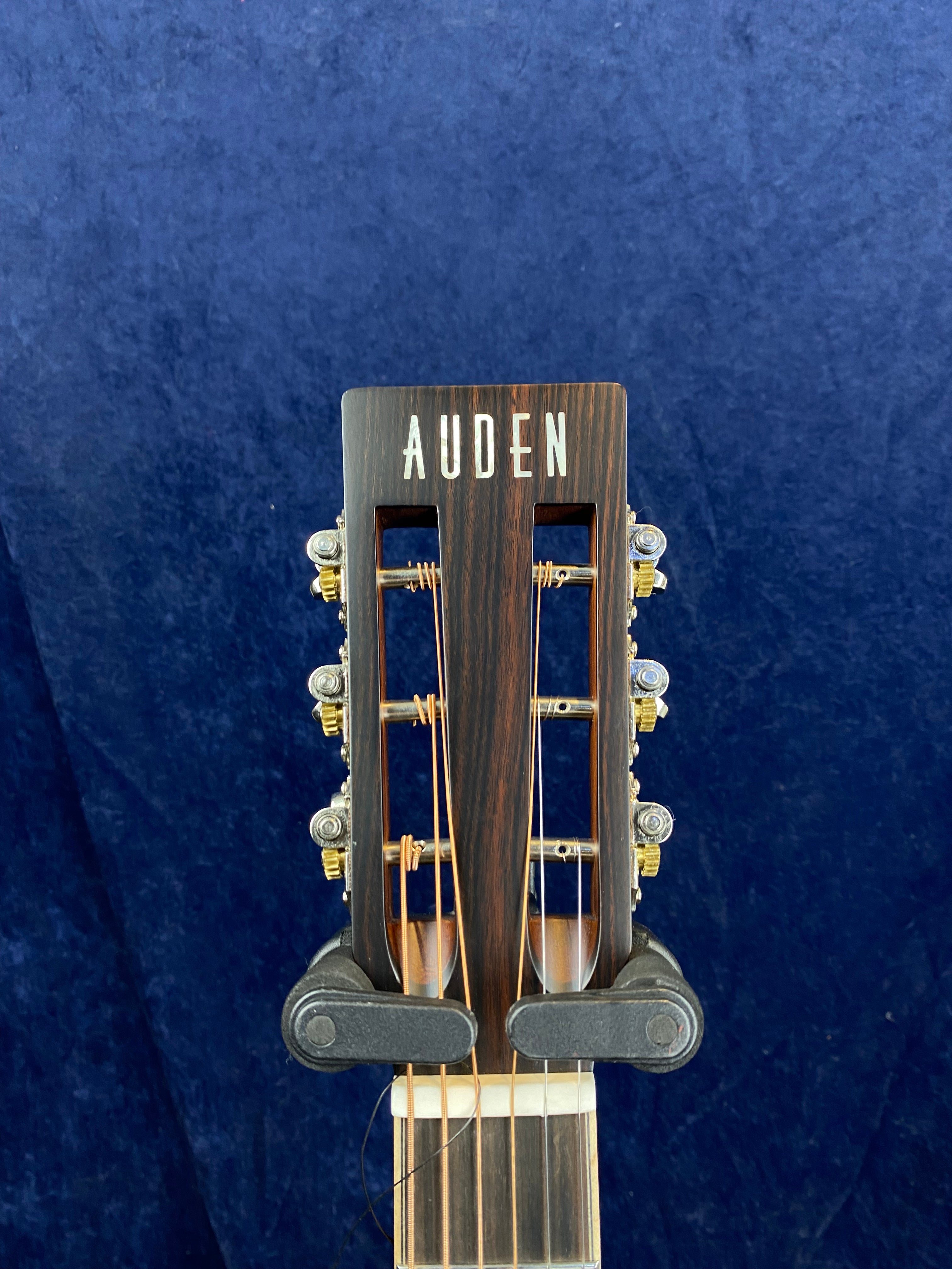 Close-up of an Auden acoustic guitar headstock with six tuning pegs, metal strings, and a dark wood finish against a dark blue background.