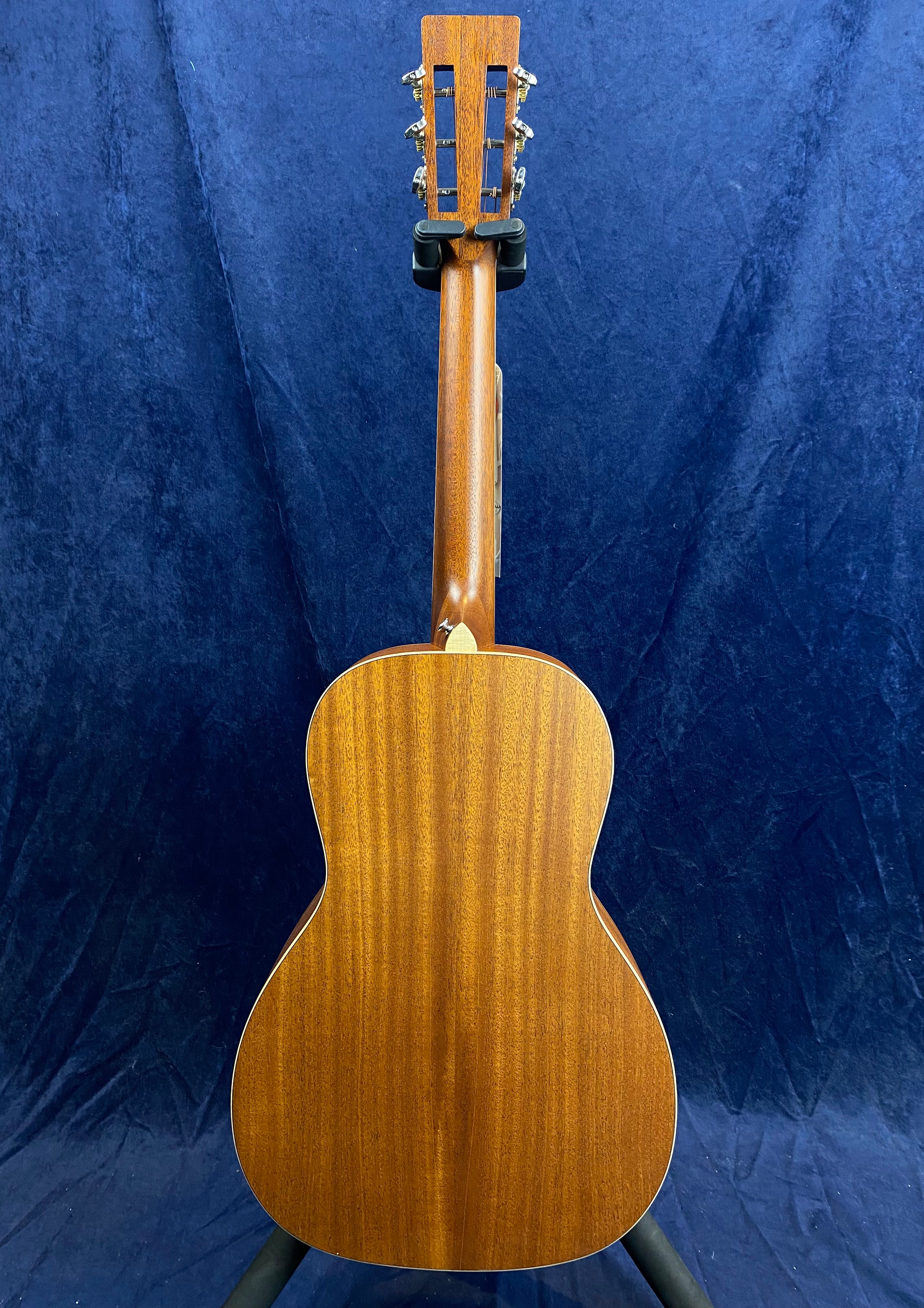 Mahogany-backed acoustic guitar facing away, showing smooth wood grain, classic shape, and slotted headstock against a dark blue background.
