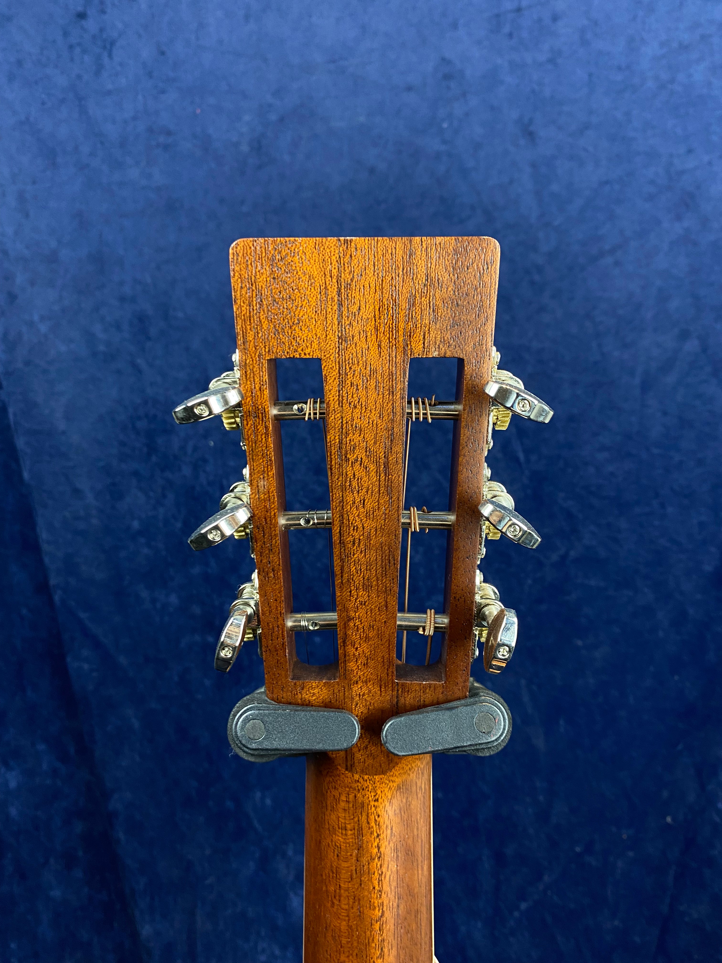 Back of an acoustic guitar headstock with open slot design and six metal tuning pegs against a dark blue background.