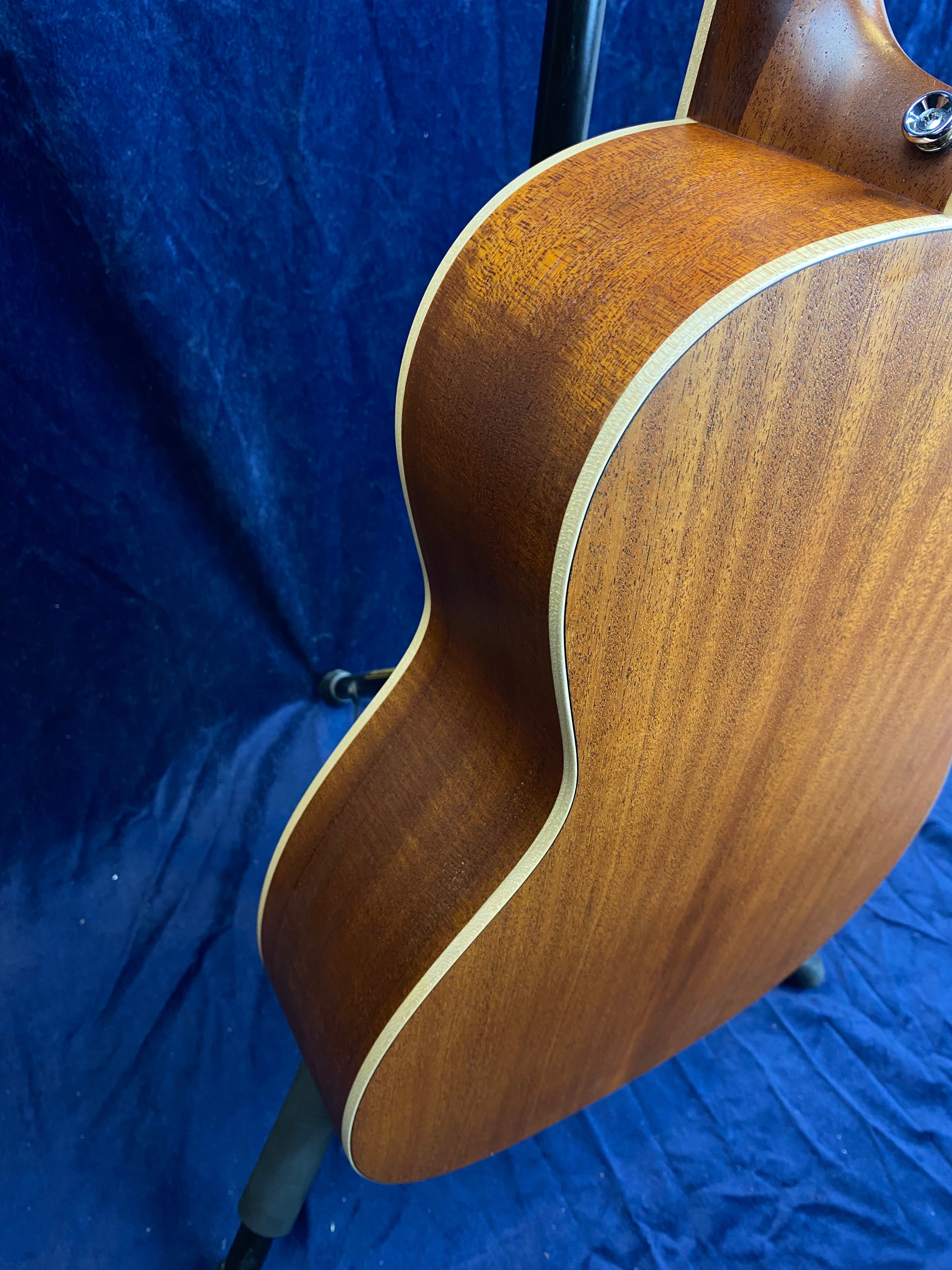 Side view of an acoustic guitar showing smooth, natural wood grain, cream edging, and part of the neck joint against a dark blue background.