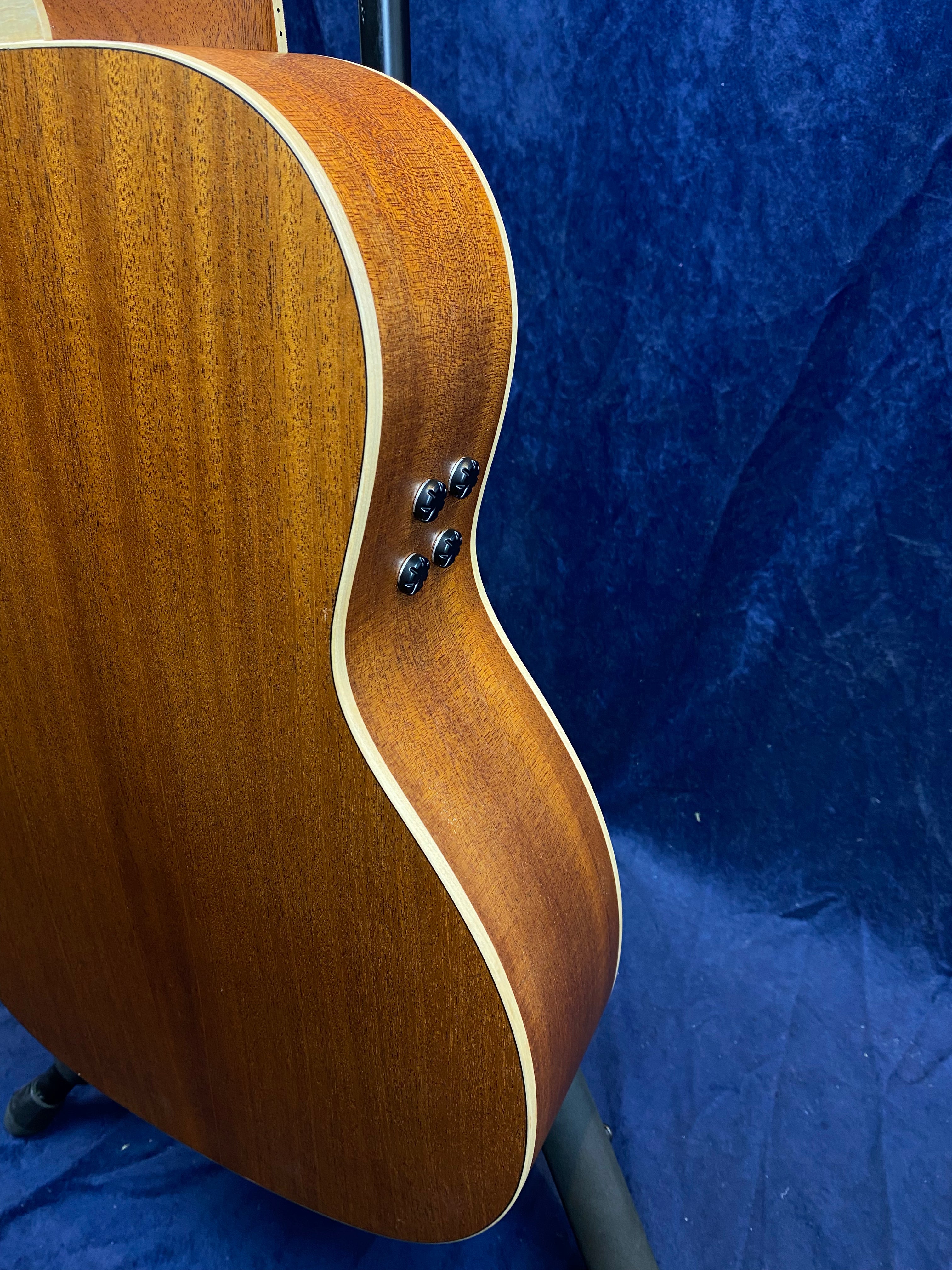 Curved side of an acoustic guitar showing wood grain, light trim, and four black control knobs on a blue background.