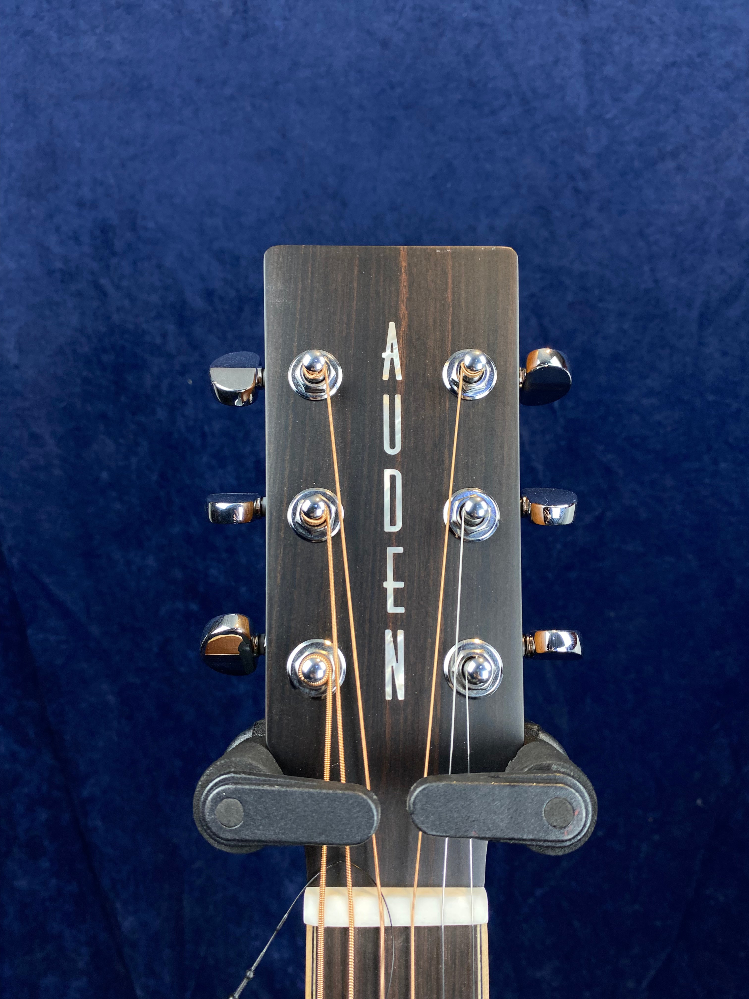 Dark wood acoustic guitar headstock with "AUDEN" in white letters, six silver tuning pegs, and a black capo clamped below the nut.