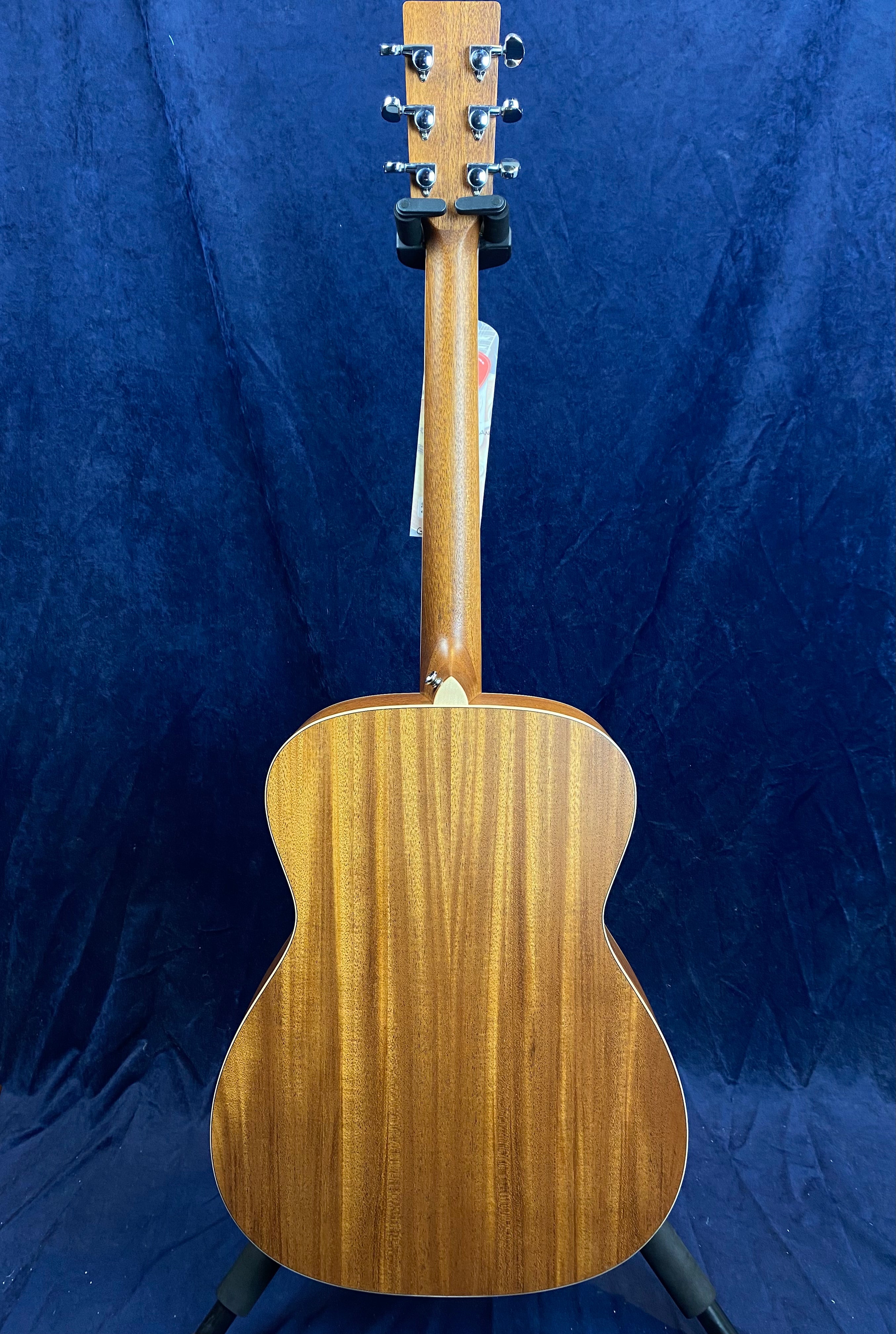 Back view of an acoustic guitar showing natural wood grain, six tuning pegs, and a smooth curved body. The guitar leans on a stand.