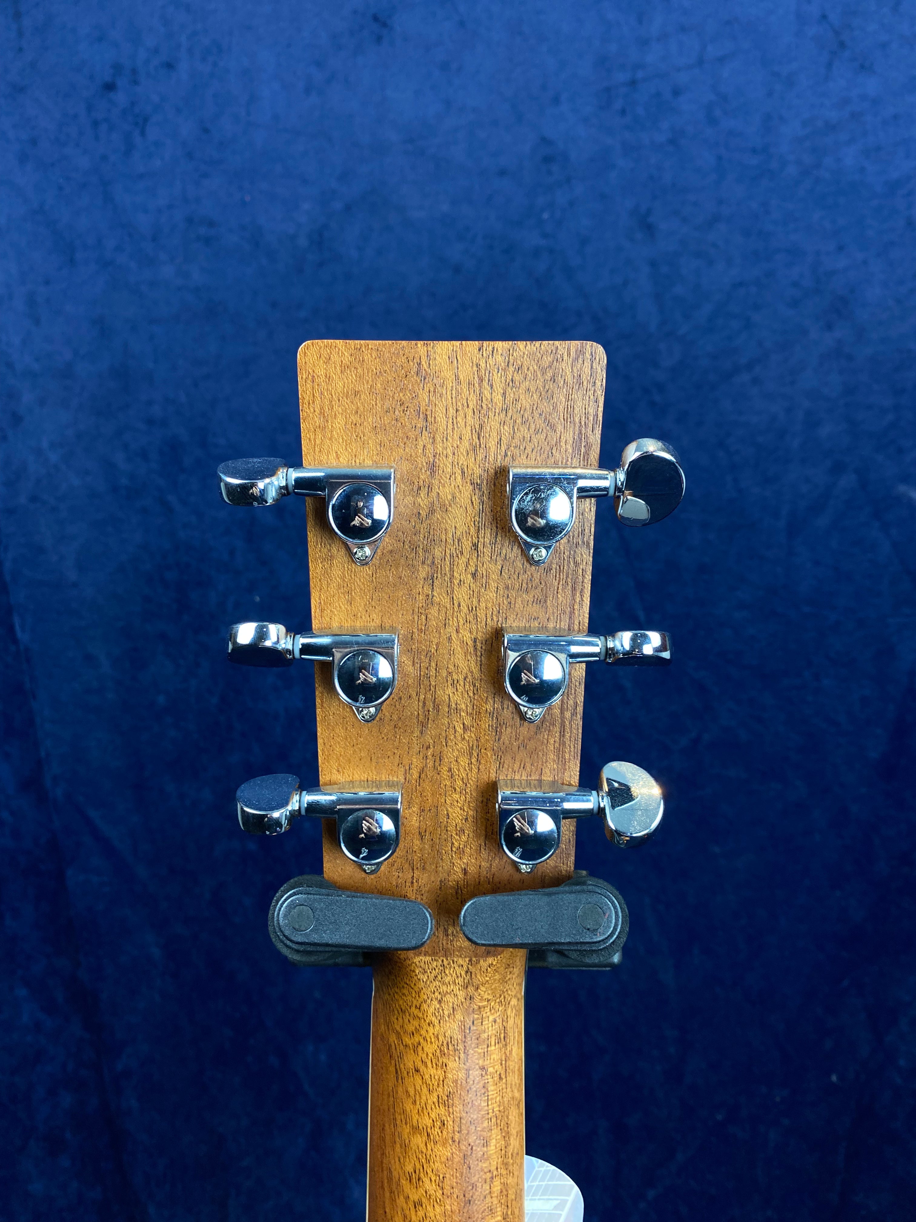 Six chrome tuning pegs on the back of a wooden acoustic guitar headstock, set against a dark blue textured background.