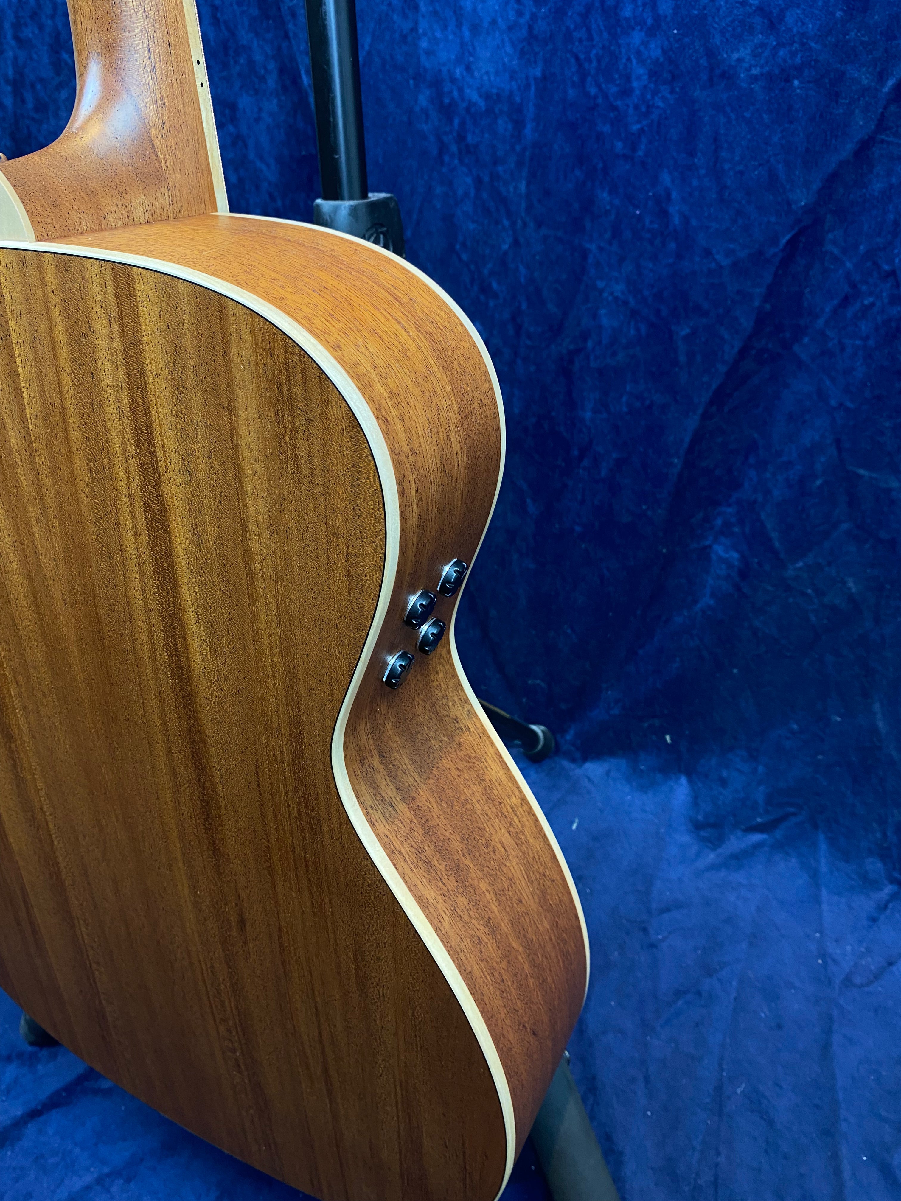 Side view of a wooden acoustic guitar showing its smooth brown back, cream binding, and two black strap buttons. Blue fabric background.