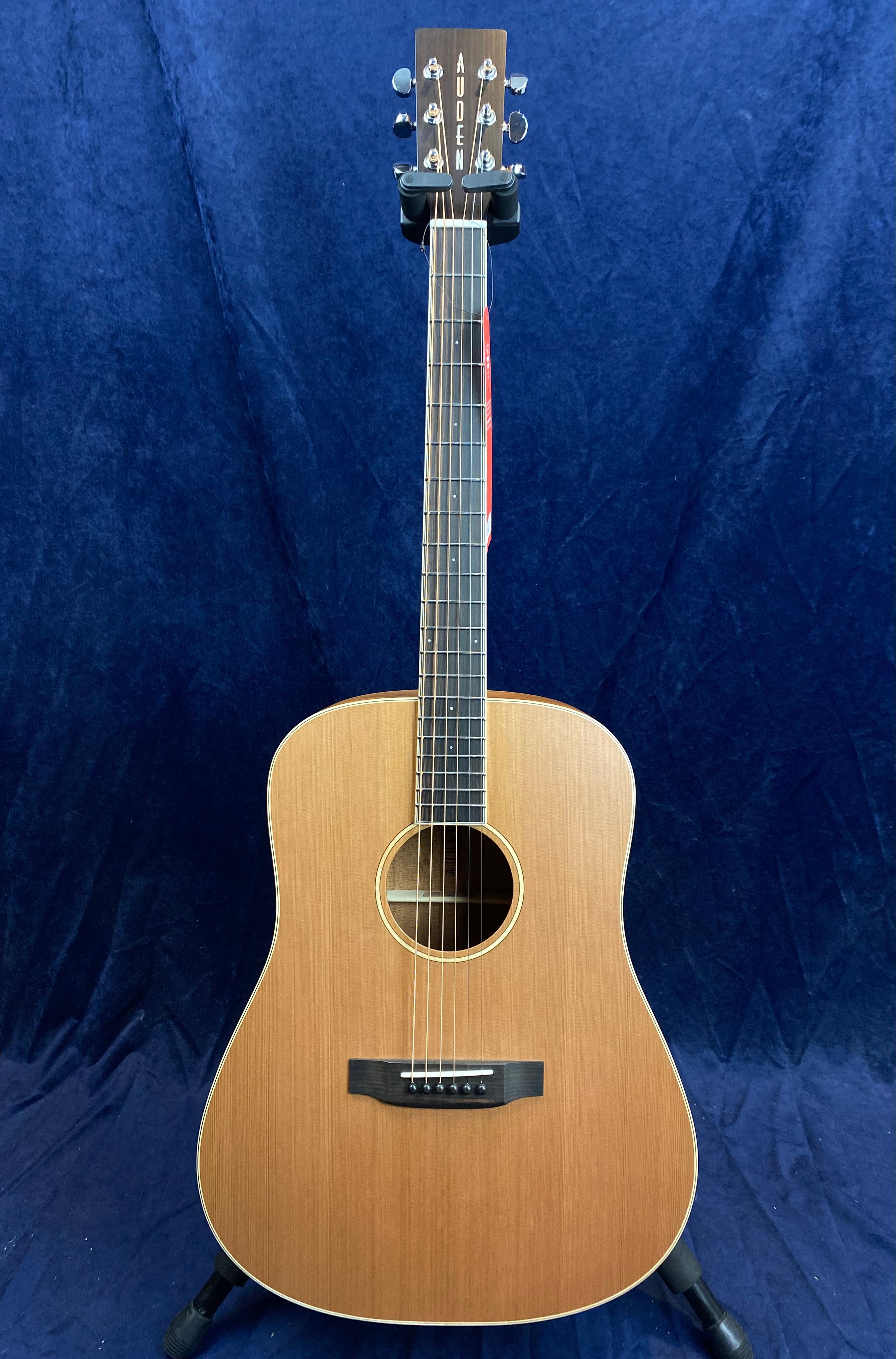 Natural wood finish acoustic guitar with a rounded body, six strings, and the brand name Auden on the headstock, standing against a dark blue background.