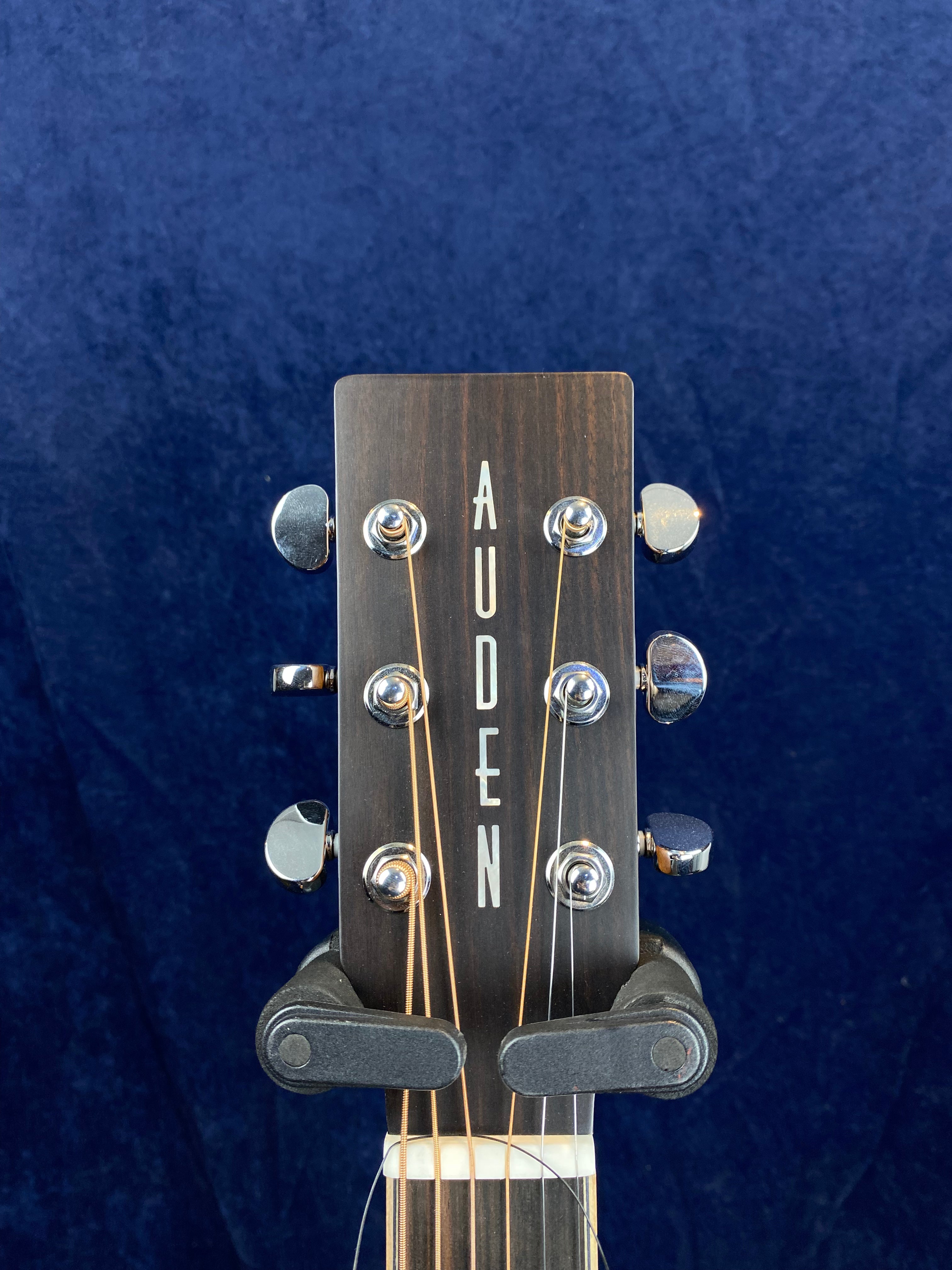 Close-up of an Auden acoustic guitar headstock with six tuning pegs, strings attached, against a plain dark blue background.