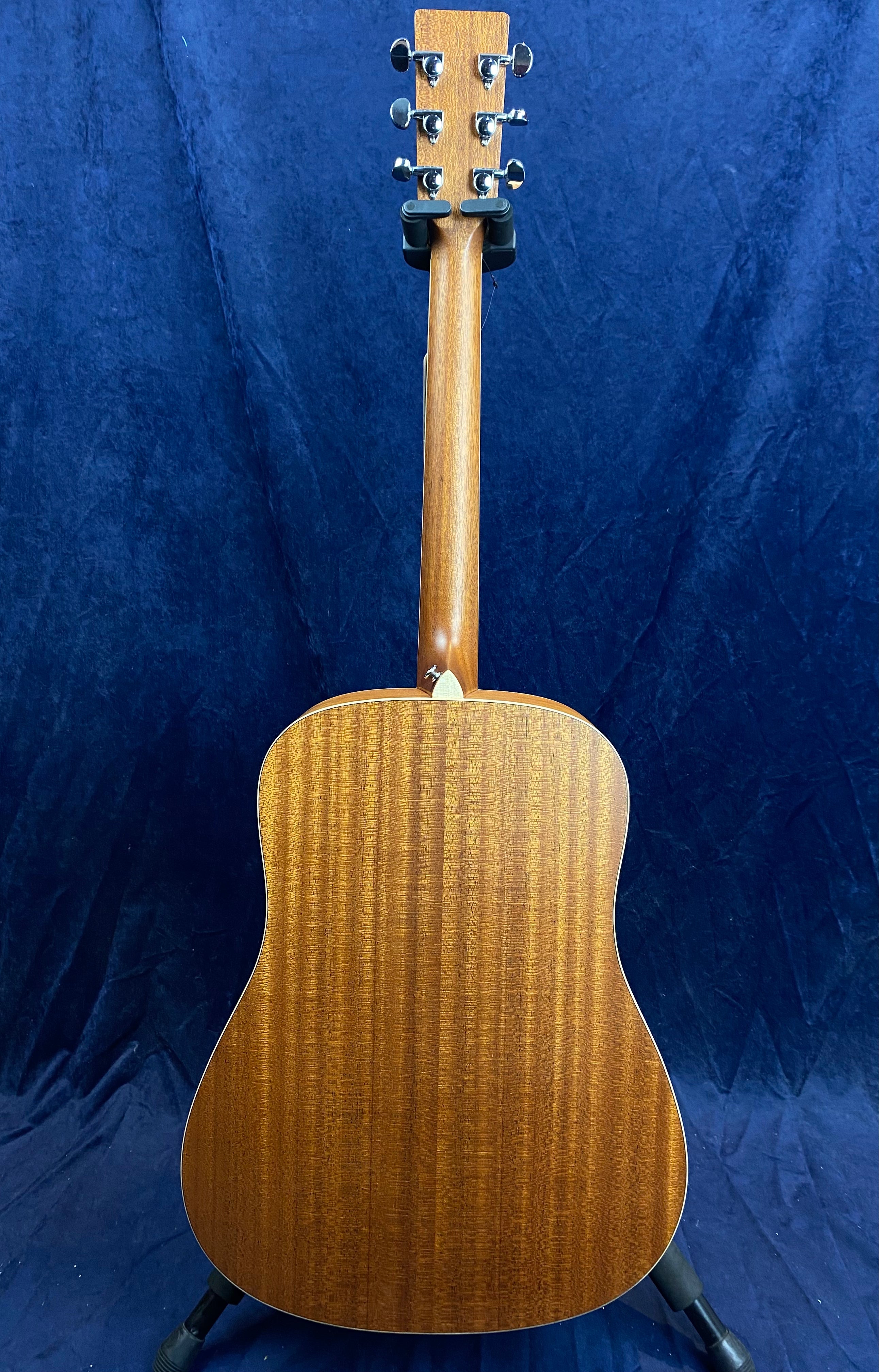 Mahogany acoustic guitar shown from the back, displaying its wooden grain pattern, six tuning pegs, and a natural brown finish against a dark blue background.