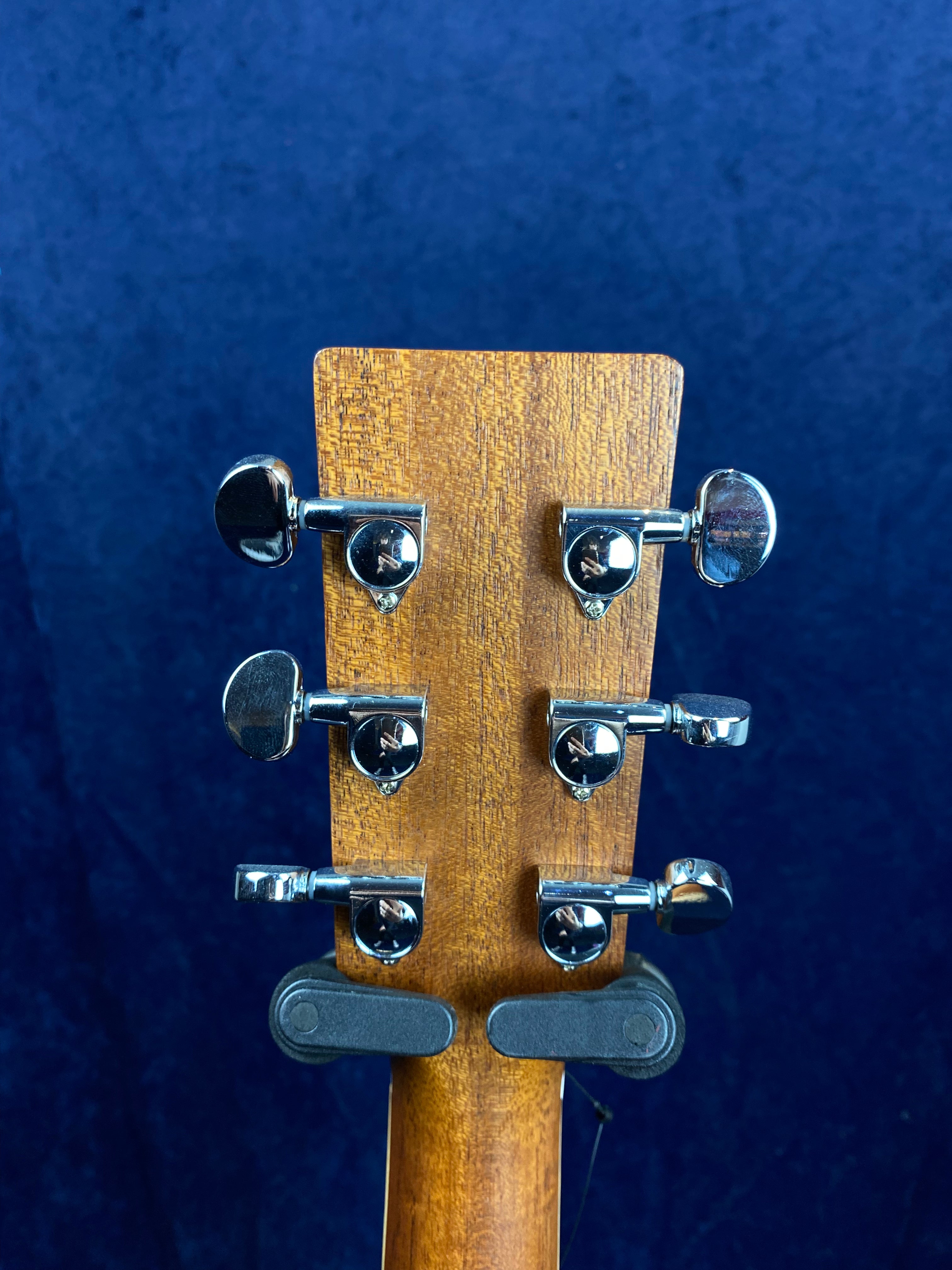 Six chrome tuning pegs on the back of a wooden acoustic guitar headstock, shown against a dark blue background.