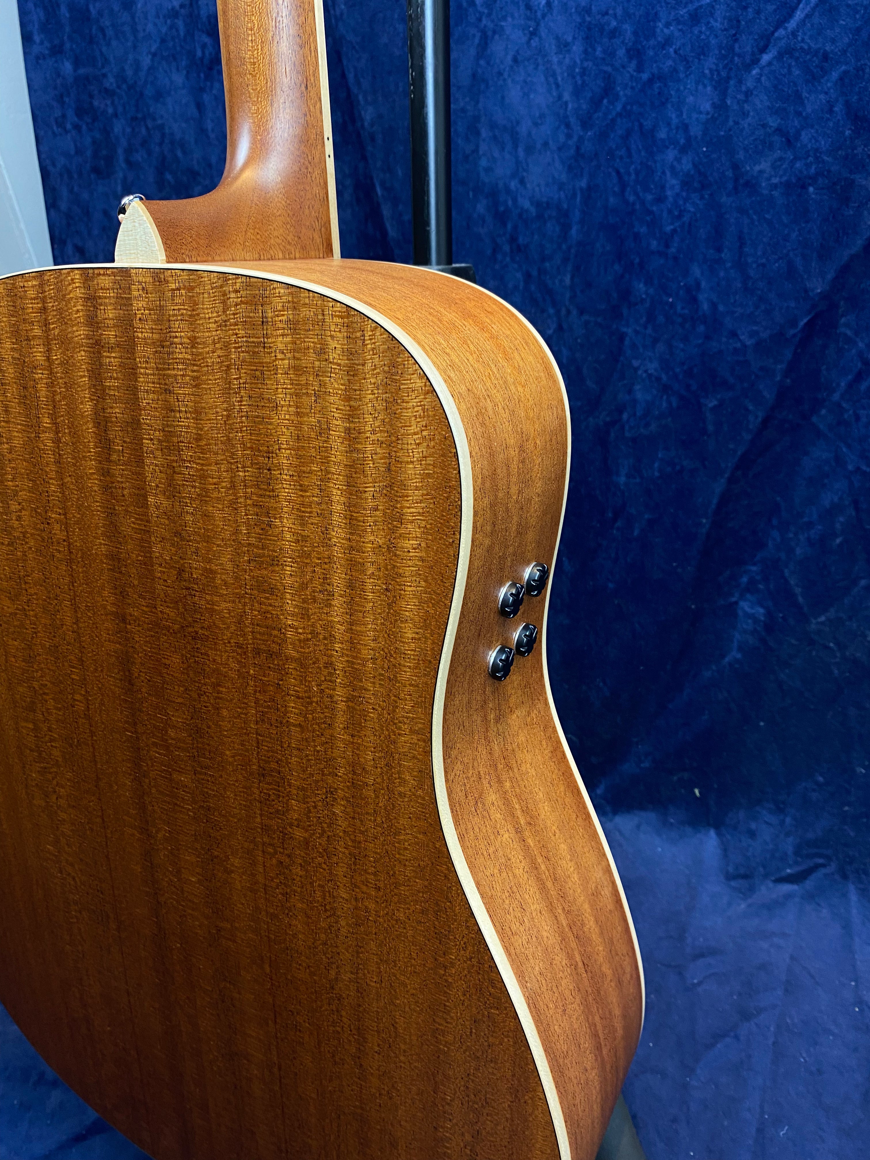 Curved mahogany back and side of an acoustic guitar with cream binding and four black control knobs on the side, set against a dark blue background.