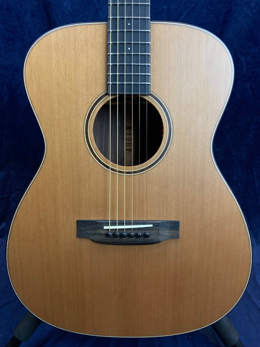 Steel-string acoustic guitar with a natural wood finish, round sound hole, black bridge, six bridge pins, and dark fretboard against a dark background.