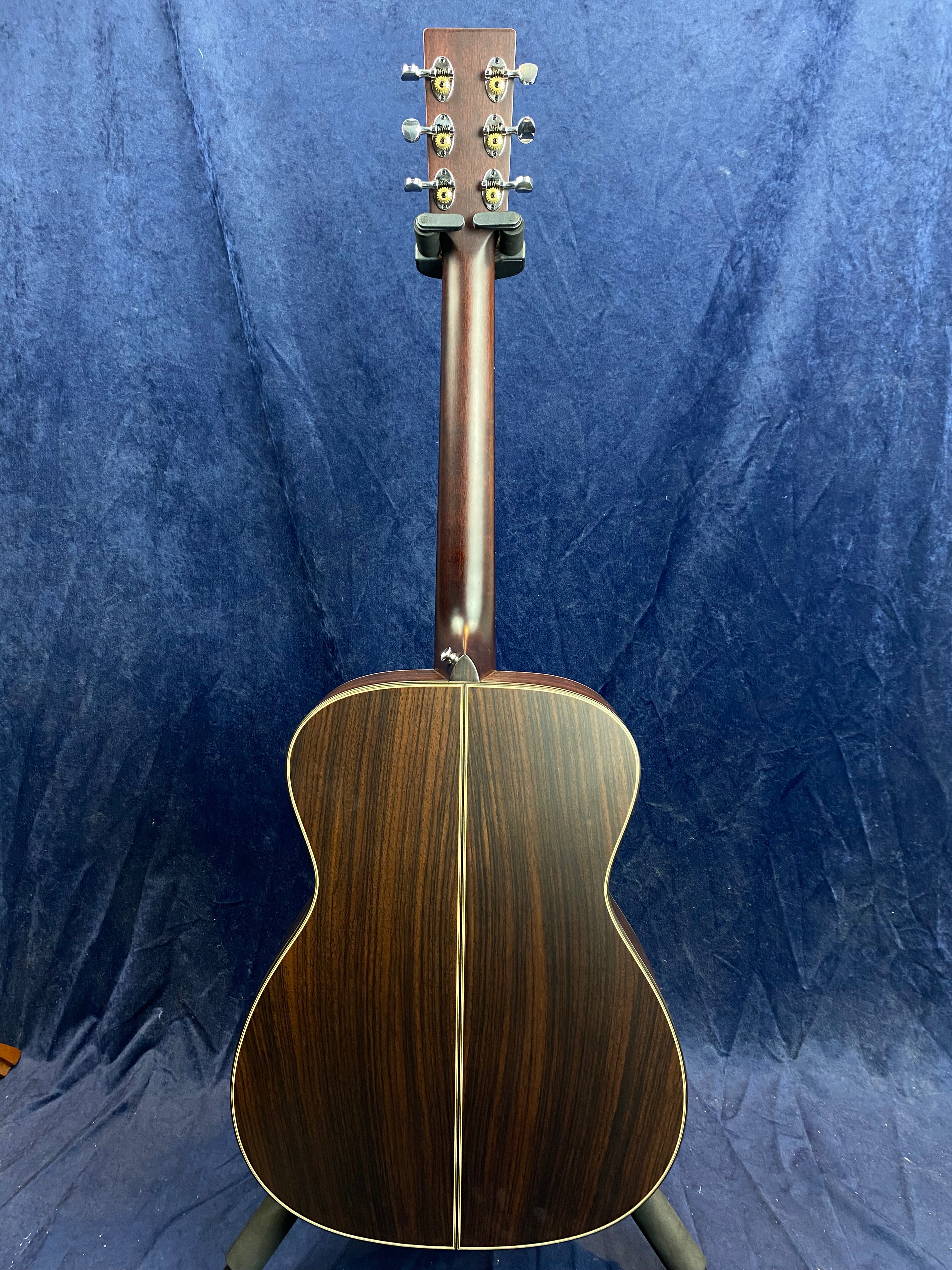 The back of an acoustic guitar with dark wood grain, ivory binding, and gold tuning pegs, shown upright against a blue background.