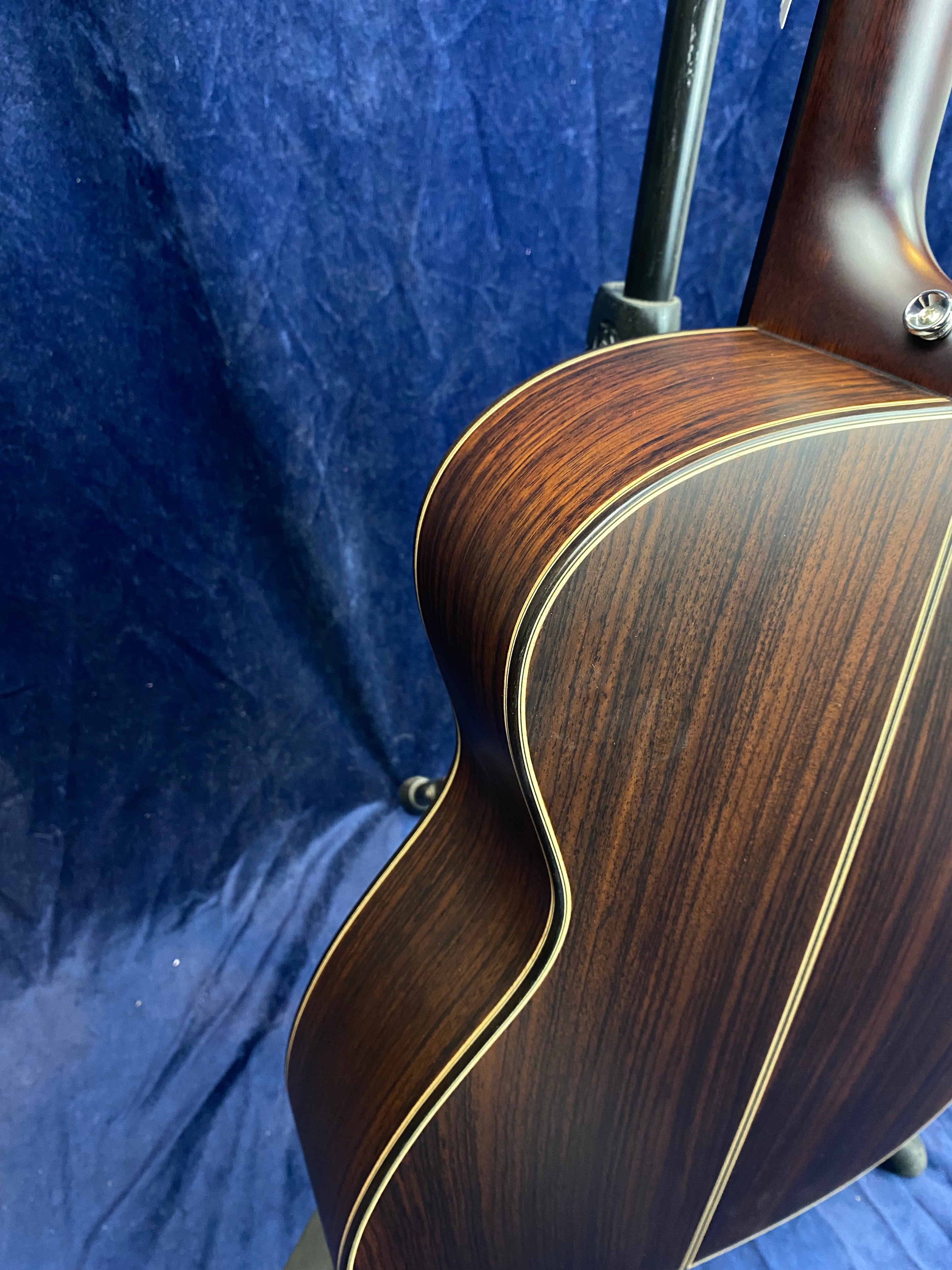 Acoustic guitar with dark wood back and sides, highlighted by light-colored binding and a central stripe, seen against a dark blue background.
