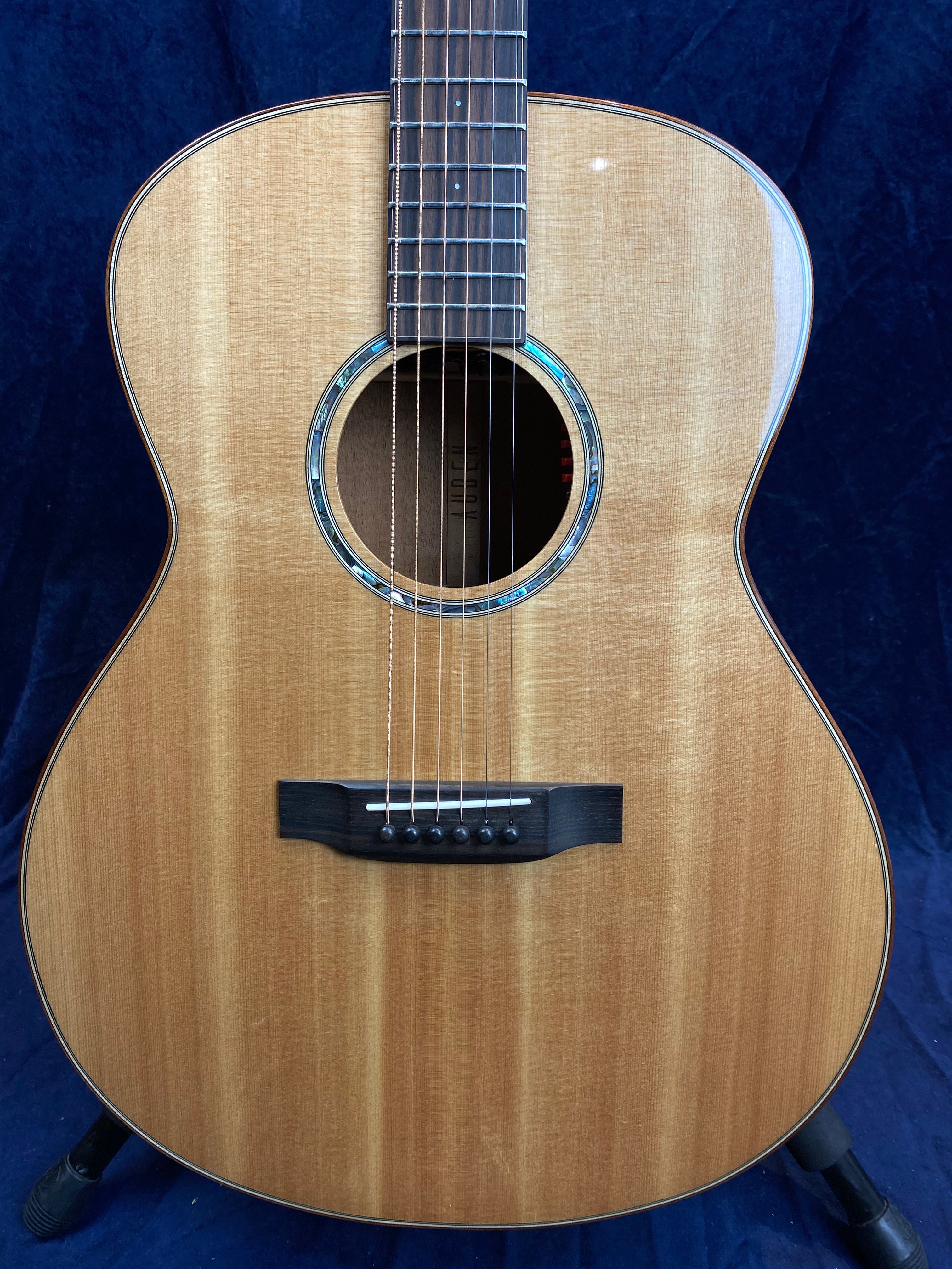 Light brown acoustic guitar with a glossy wood finish, a round sound hole bordered by abalone inlay, six strings, and a dark wooden bridge with white bridge pins.