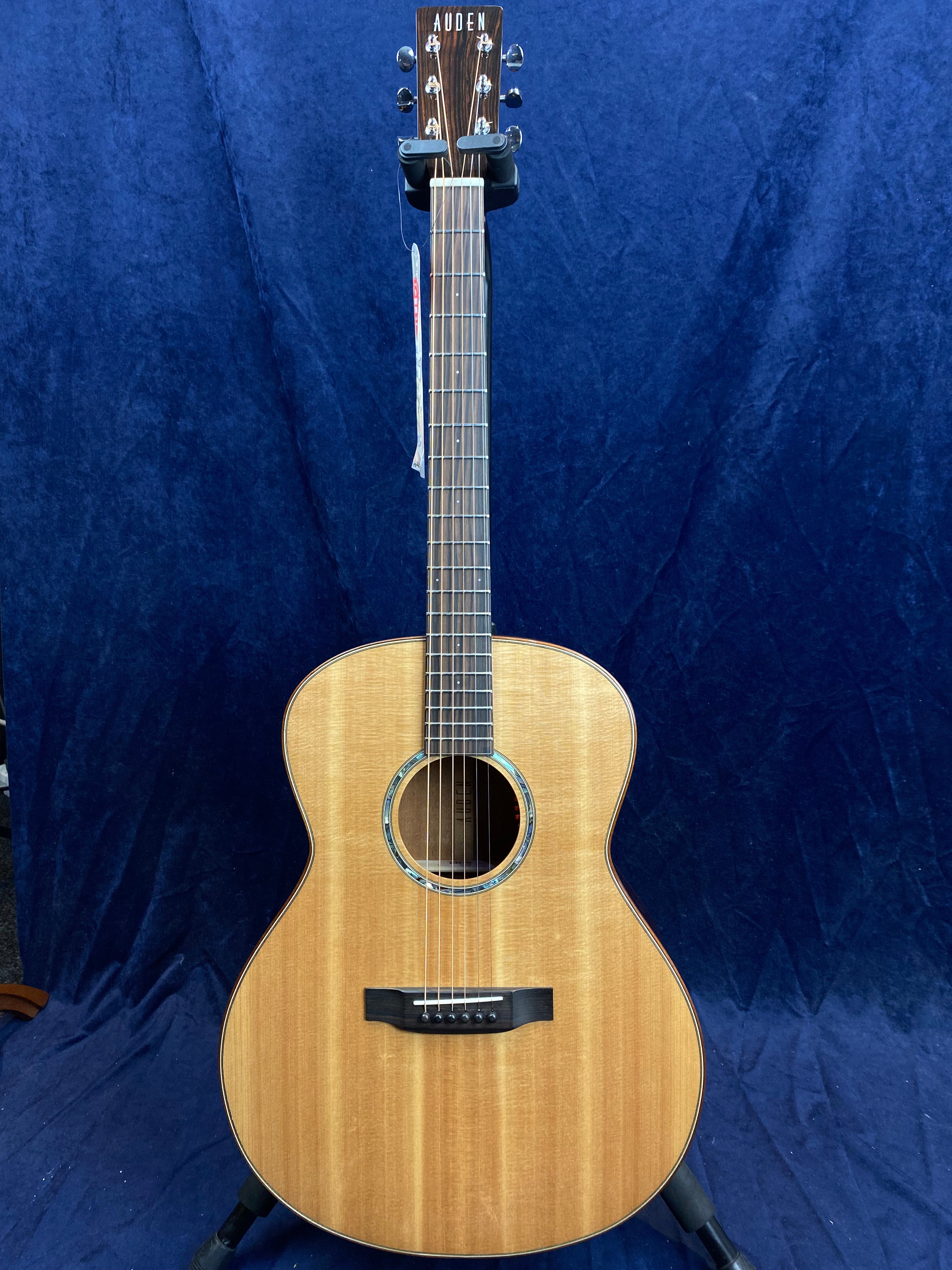 Natural wood Auden acoustic guitar with six strings, a round sound hole trimmed in abalone, and a dark wood bridge, displayed upright against a blue background.