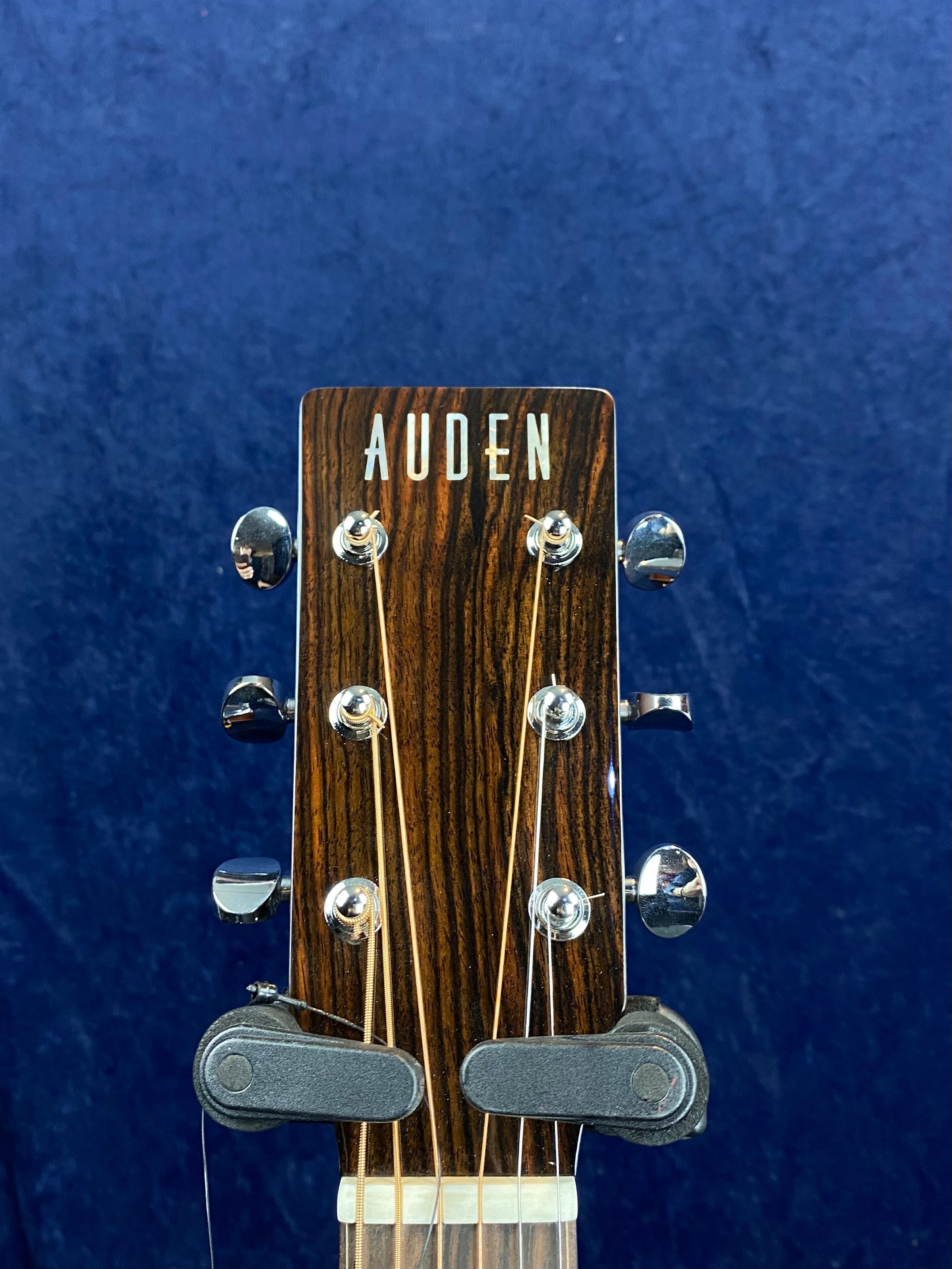 Auden acoustic guitar headstock with six chrome tuning pegs, wood grain finish, and string winders attached, set against a blue background.