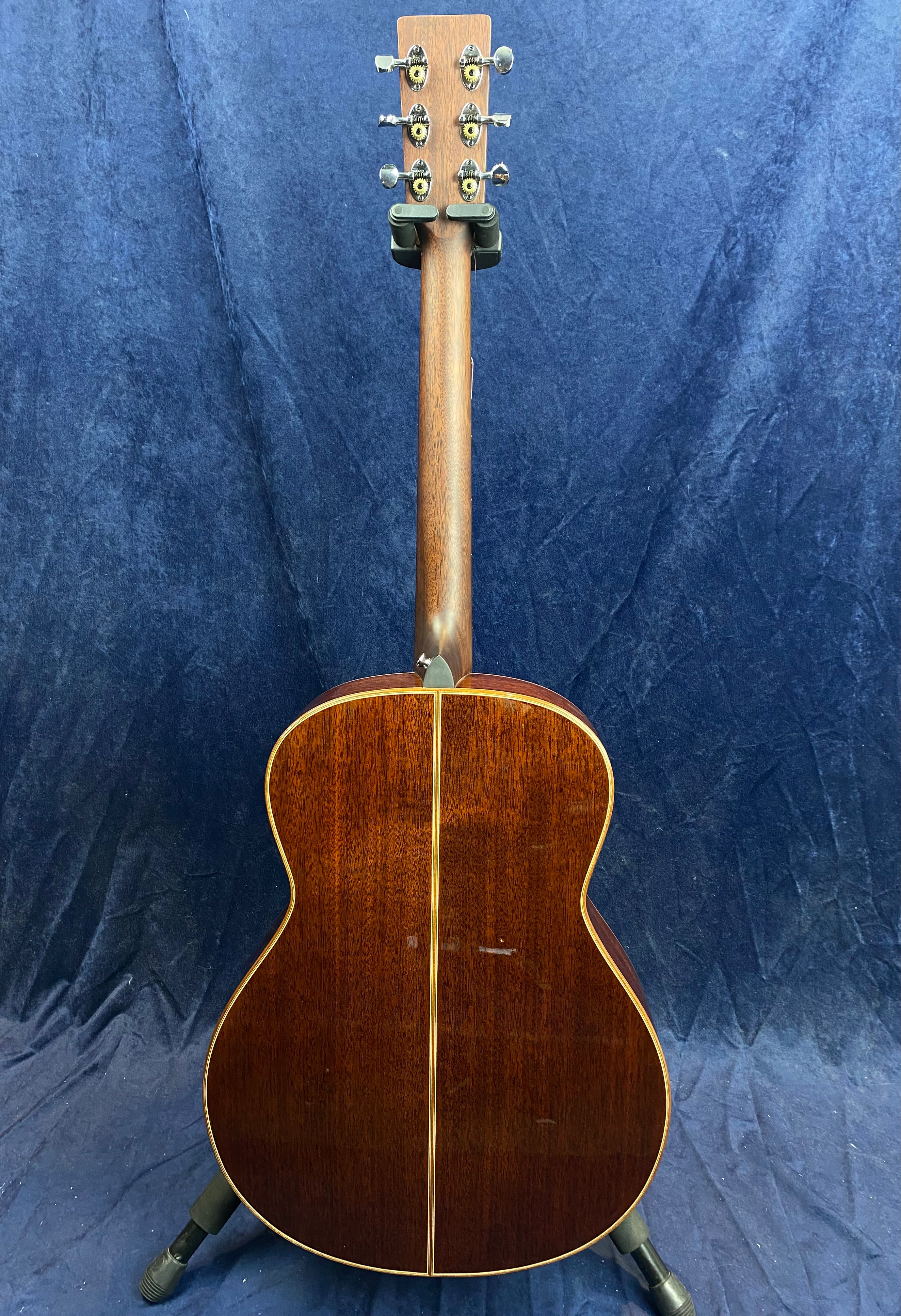 Acoustic guitar shown from the back with a glossy dark wood finish, a vertical center seam, and metal tuning pegs on the headstock.