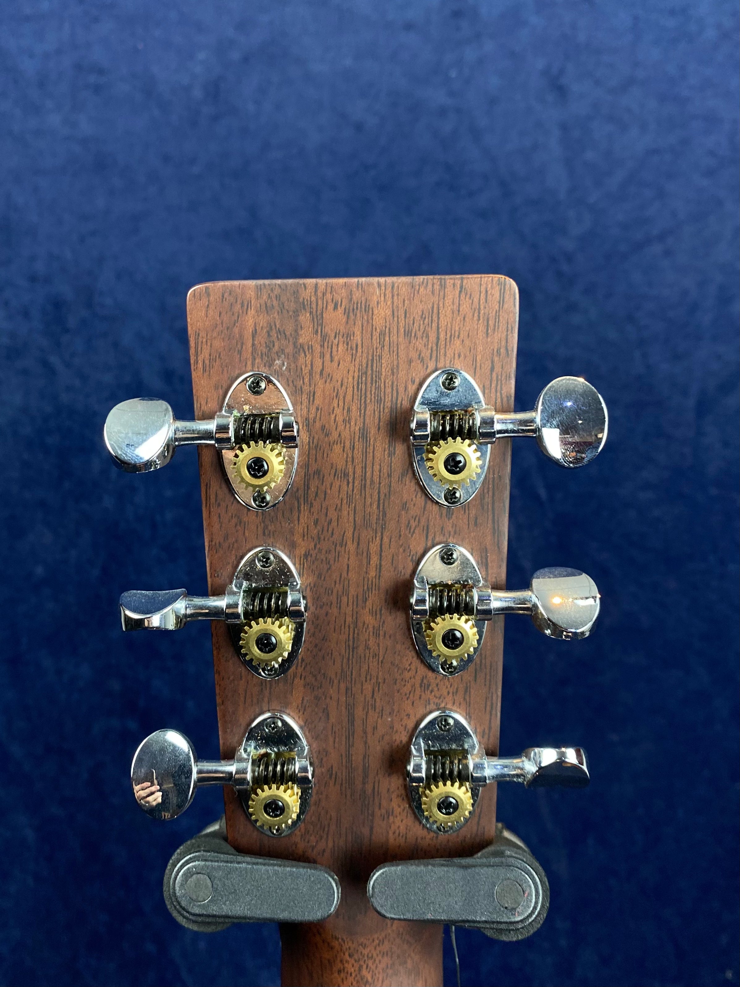 Six silver tuning pegs with exposed yellow gears are mounted on the dark wooden headstock of an acoustic guitar, against a blue background.