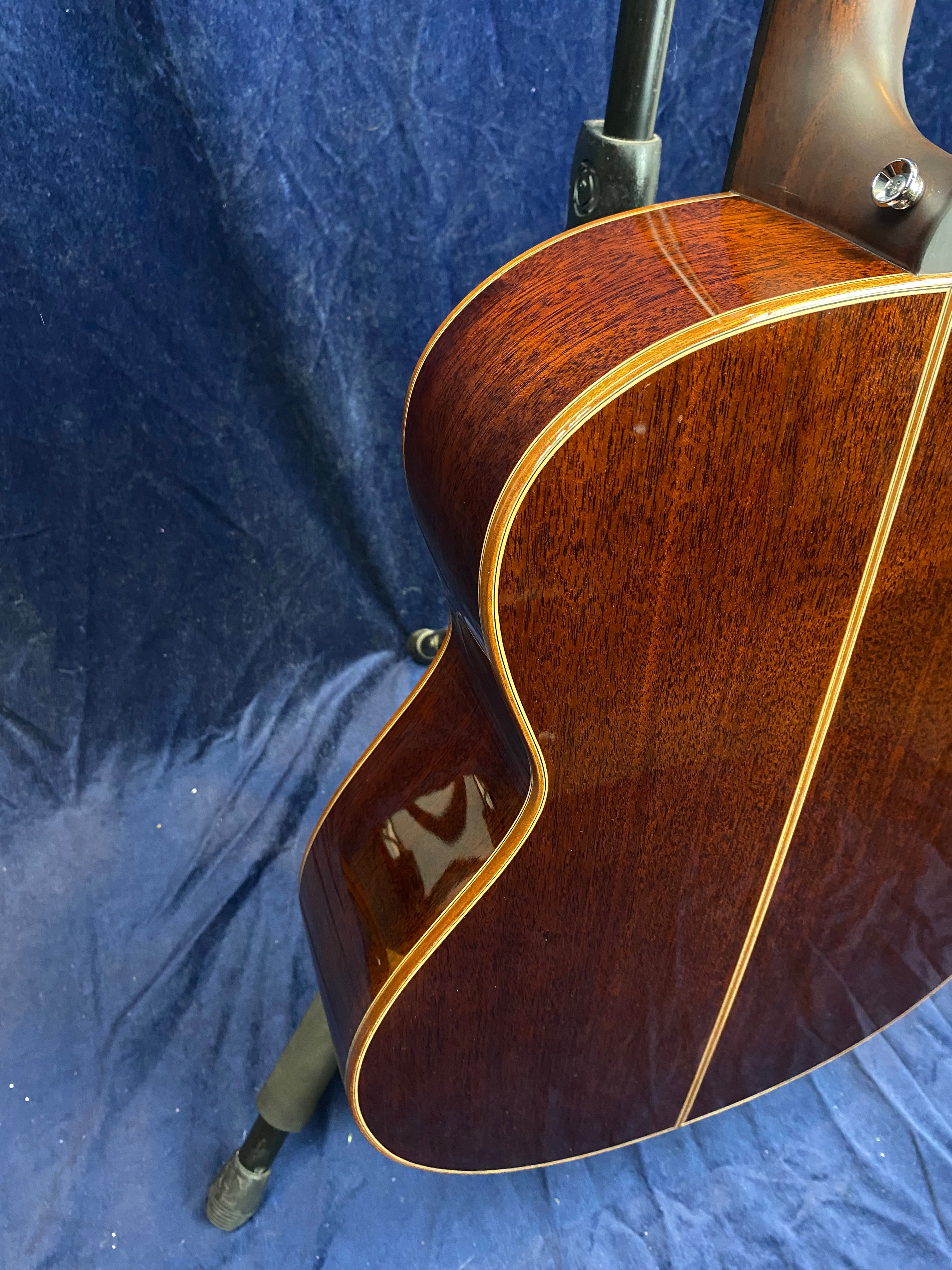 Glossy dark wood acoustic guitar body with light wood trim, shown from the side and back against a deep blue fabric background.