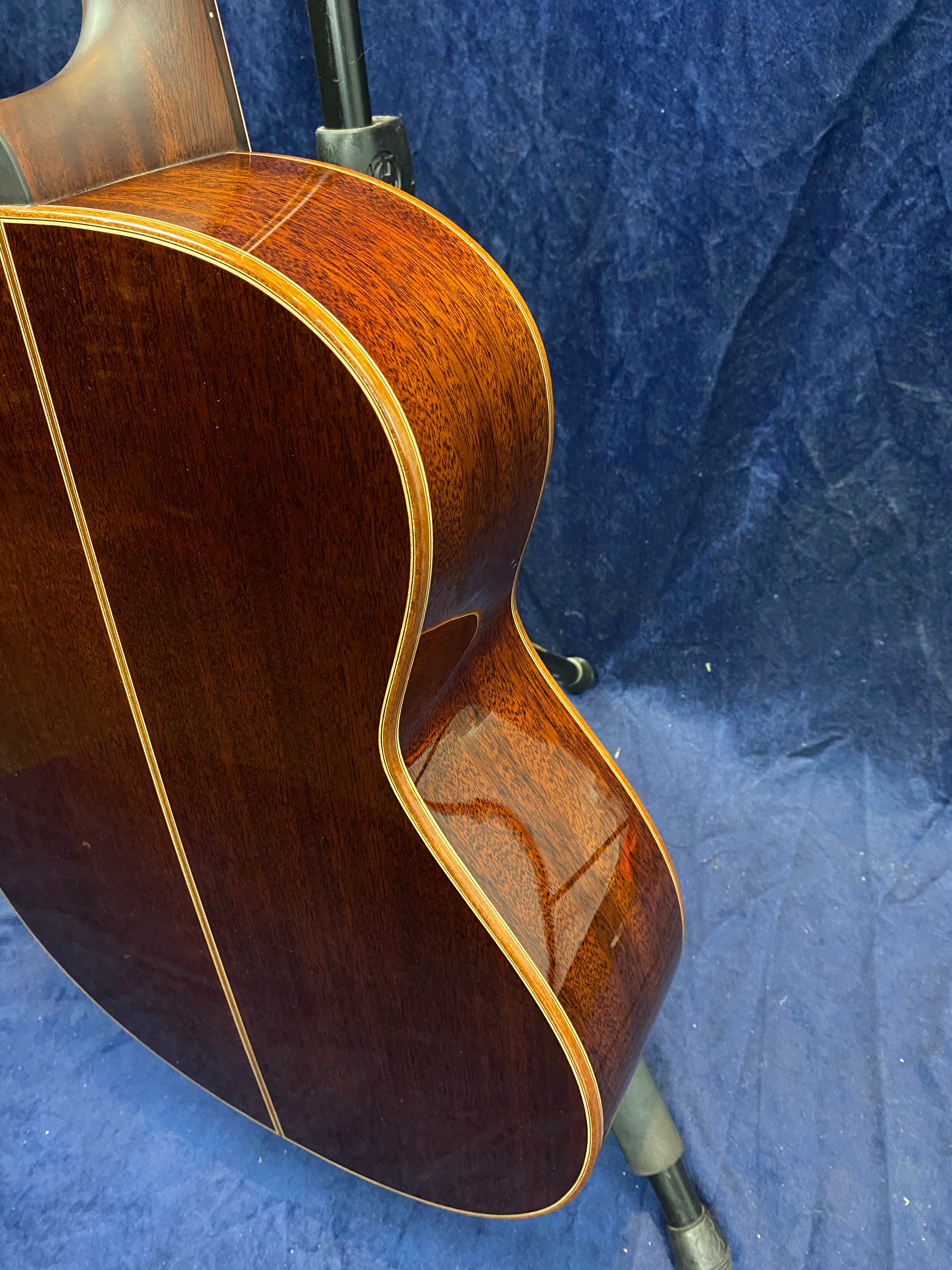 Glossy, rich brown acoustic guitar body with cream binding, resting on a stand, set against a dark blue textured background.