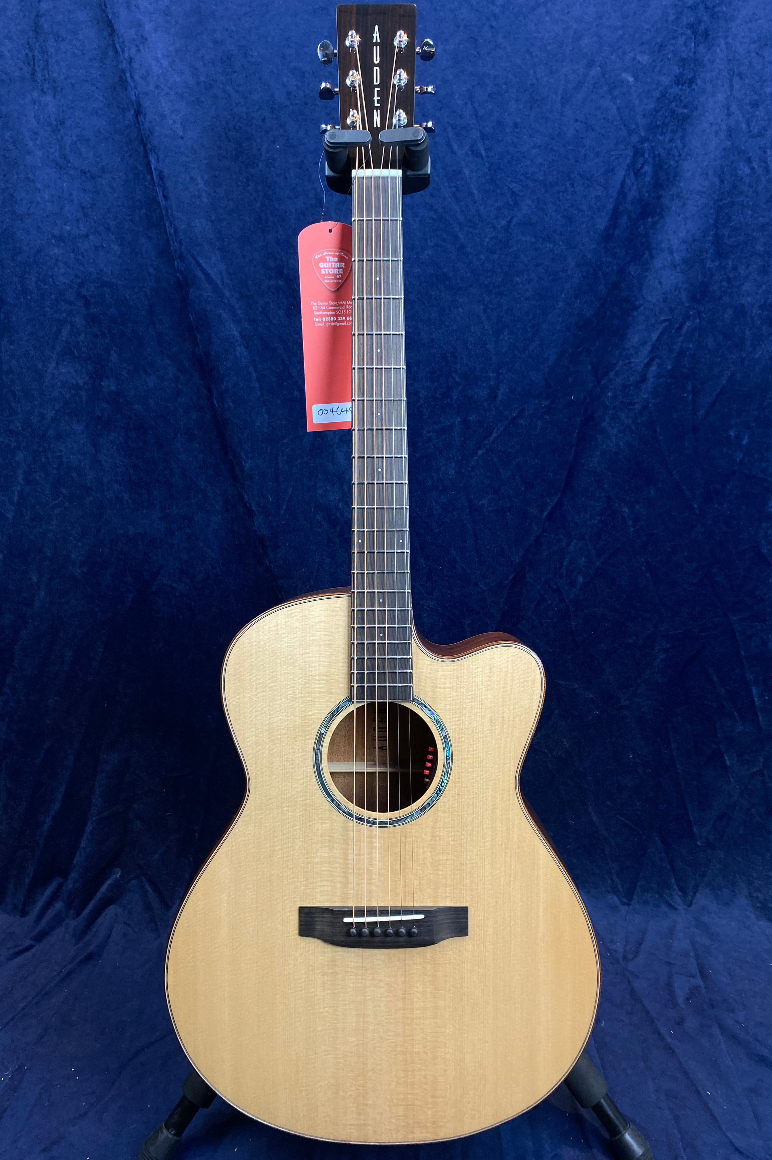 Light wood acoustic guitar with a single cutaway body, dark fretboard, and the brand name "Auden" on the headstock, displayed against dark blue fabric.