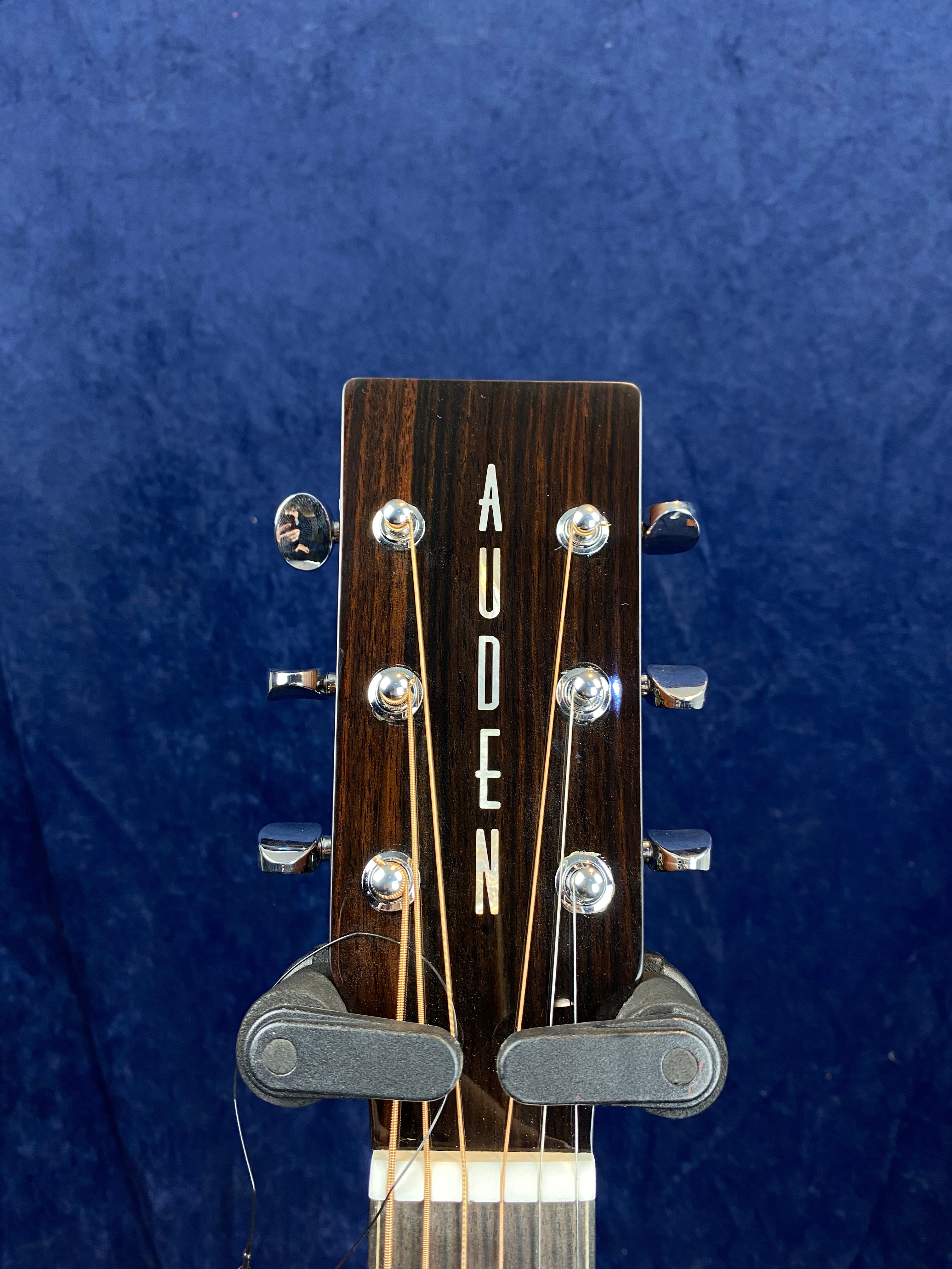 Close-up of an acoustic guitar headstock with the brand name "Auden" in white letters on dark wood, metal tuning pegs, and strings visible.