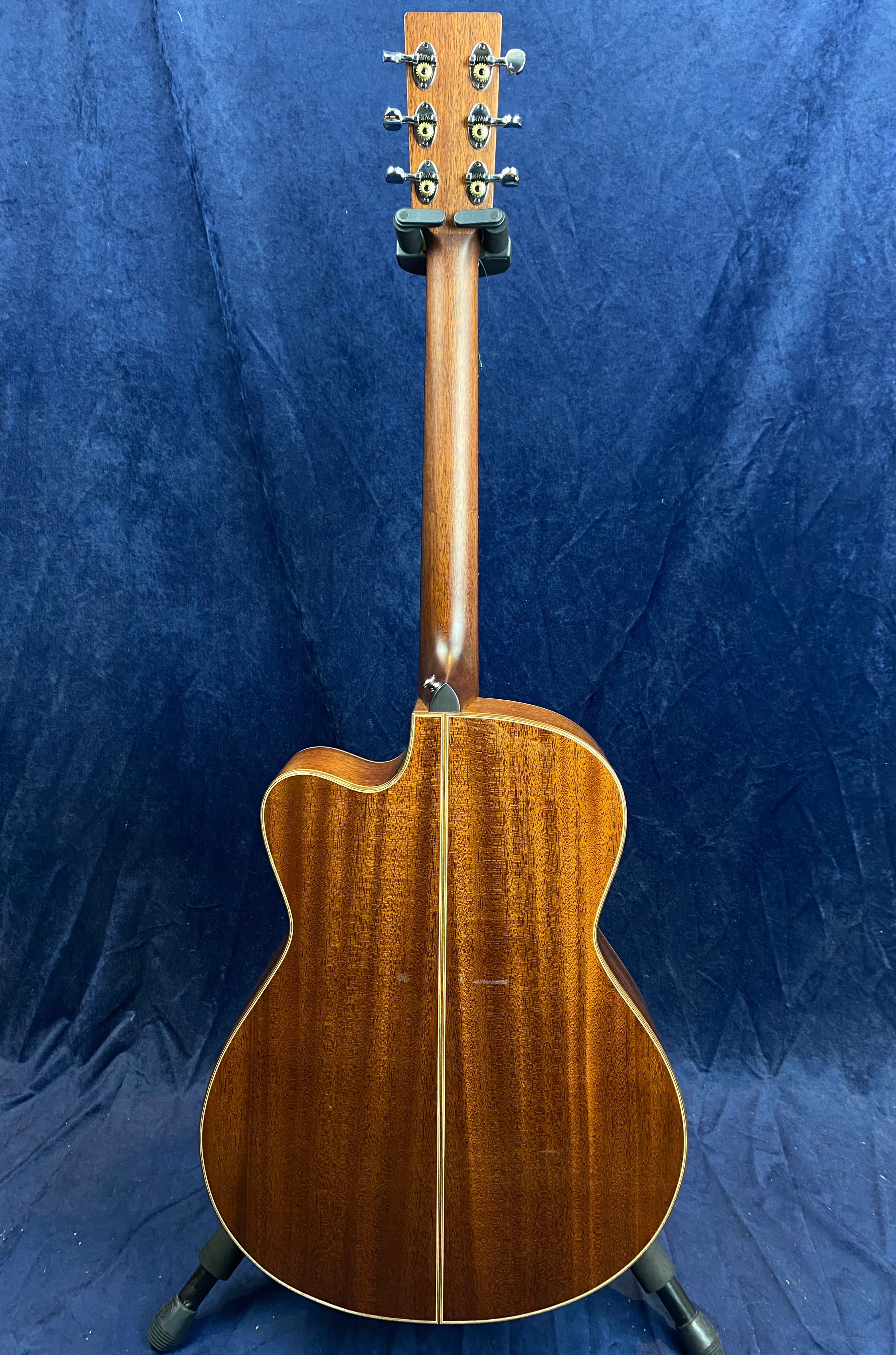 Glossy brown acoustic guitar rear view with a visible wood grain, six tuning pegs, and a single light strip running down the back’s center.