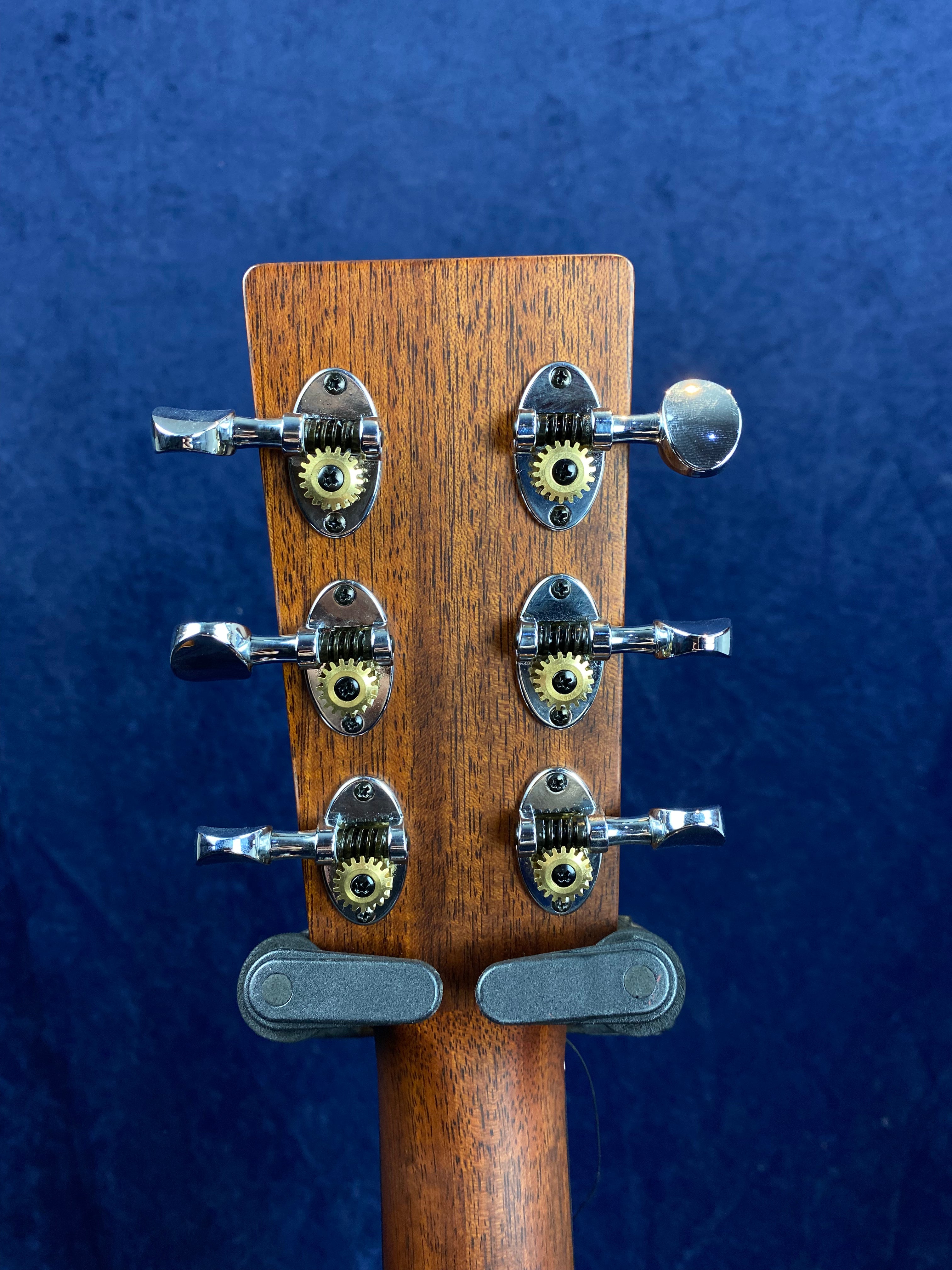 Rear view of an acoustic guitar headstock shows six chrome tuning pegs with exposed gears on a natural wood finish, set against a blue background.