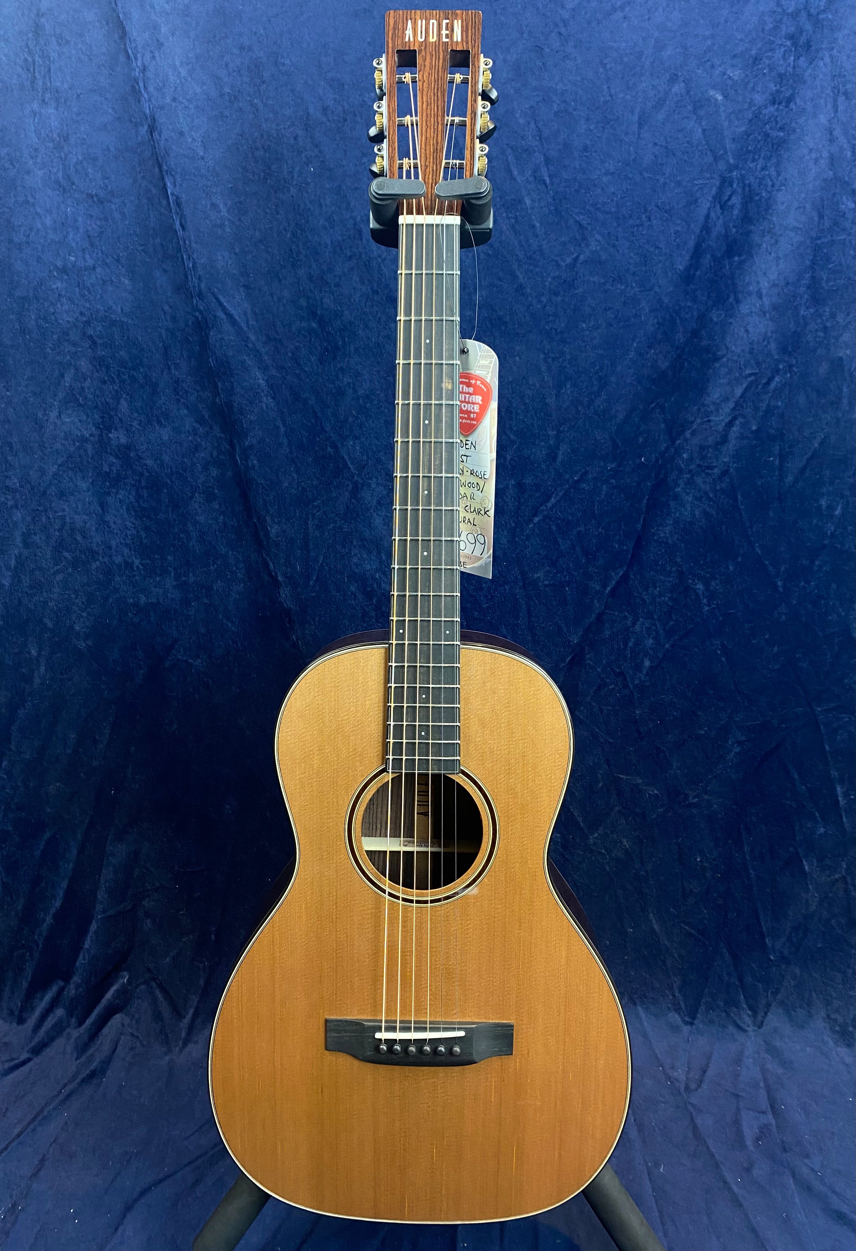 Small-bodied acoustic guitar with a natural wood finish, slotted headstock, and six strings, standing upright against a dark blue background.