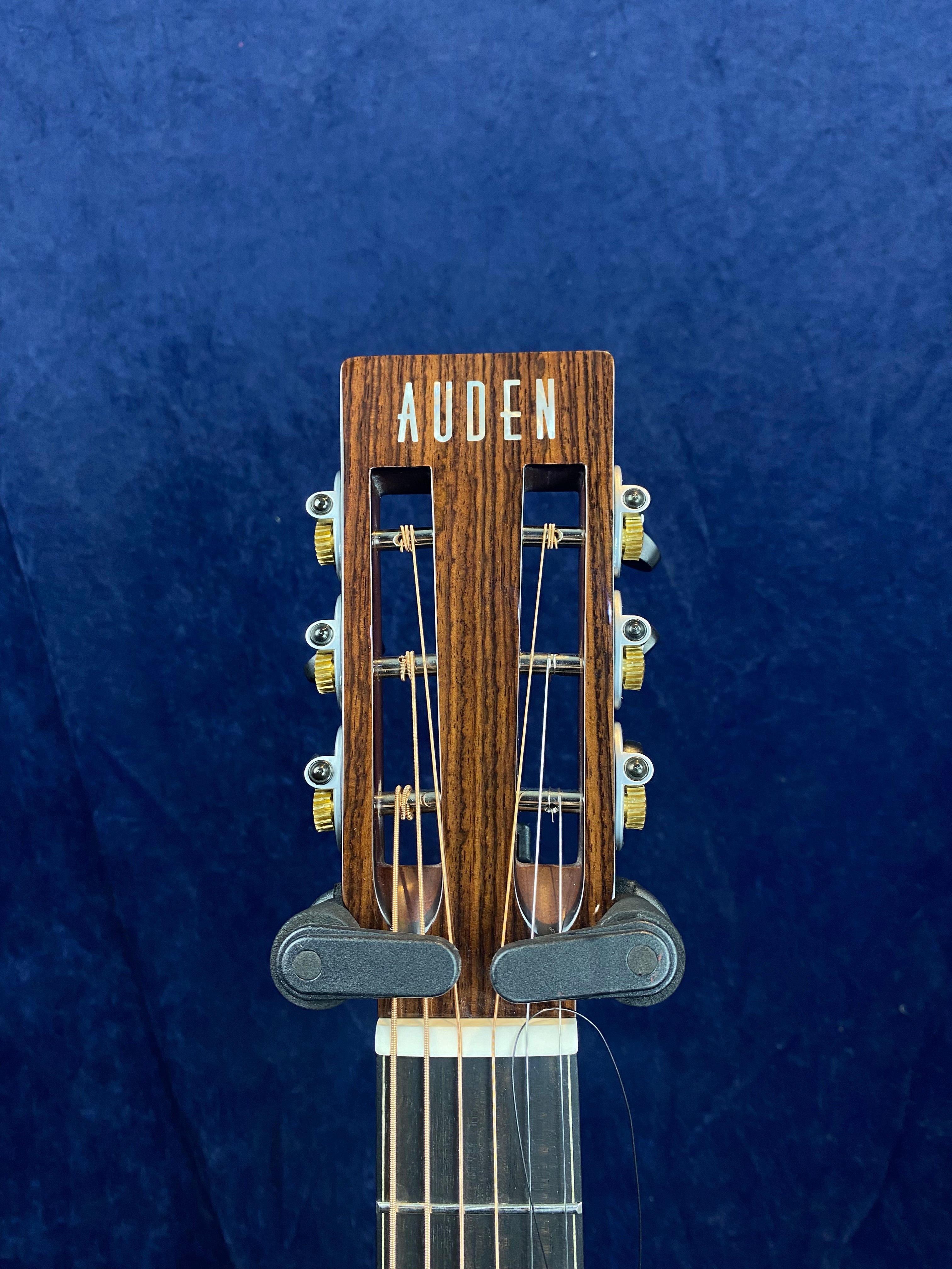 The headstock of an Auden acoustic guitar shows six tuning pegs, gold gear hardware, and dark wood grain against a blue background.