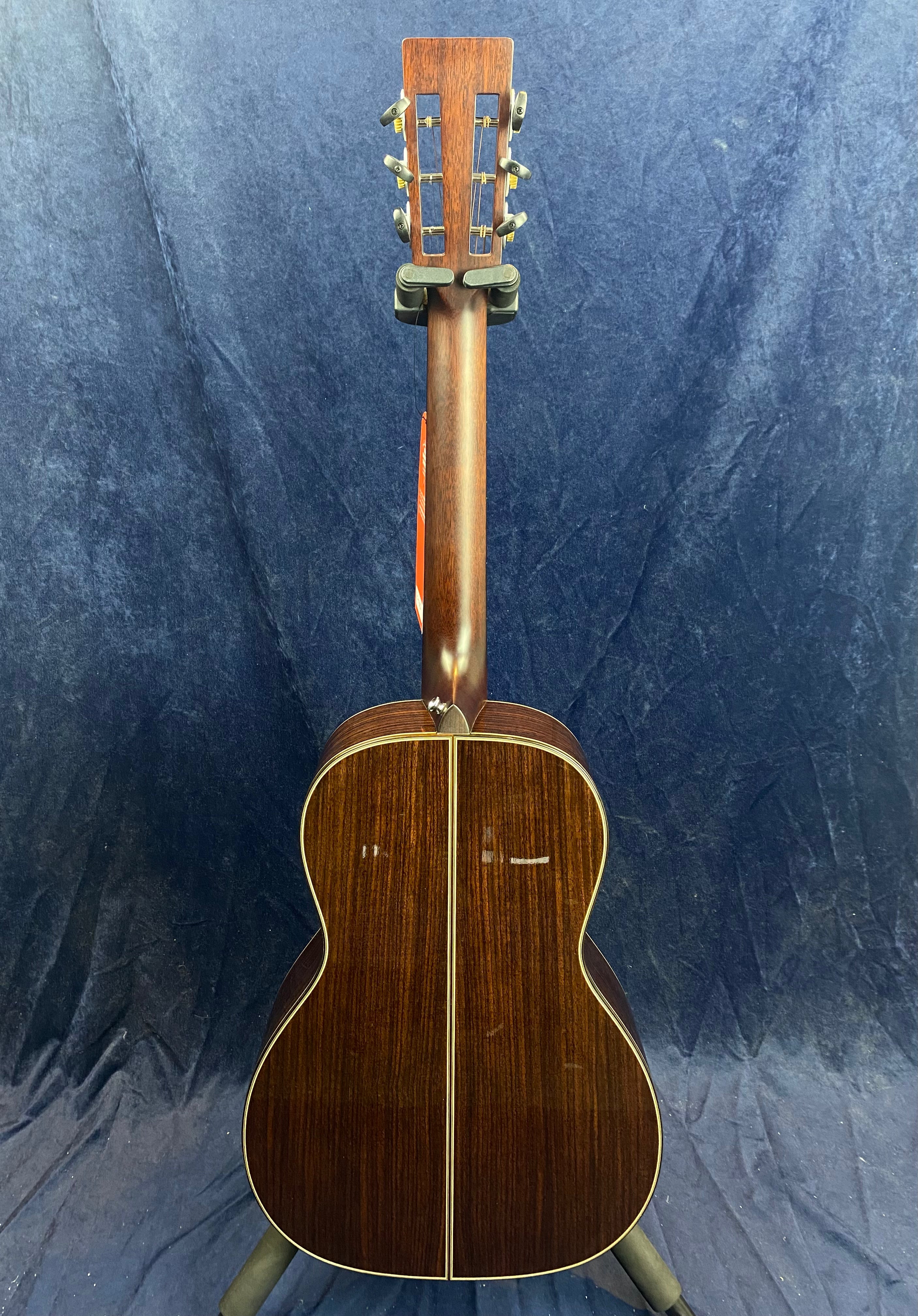 Acoustic guitar shown from the back, dark wood body with a central light wood inlay stripe, slotted headstock with tuning pegs, standing on a floor stand.