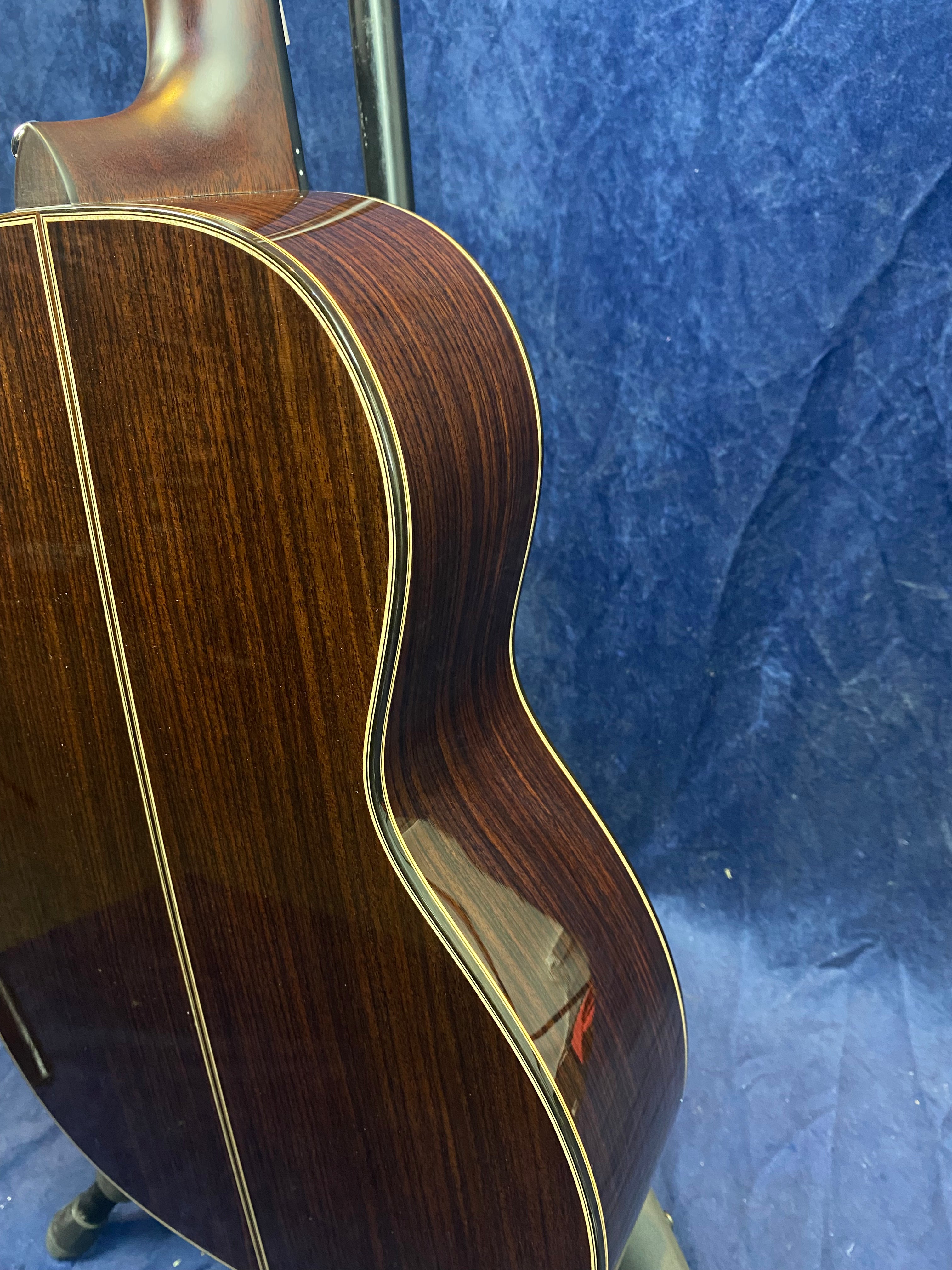 Curved side and back of an acoustic guitar made of dark polished wood with light trim, shown against a textured blue background.