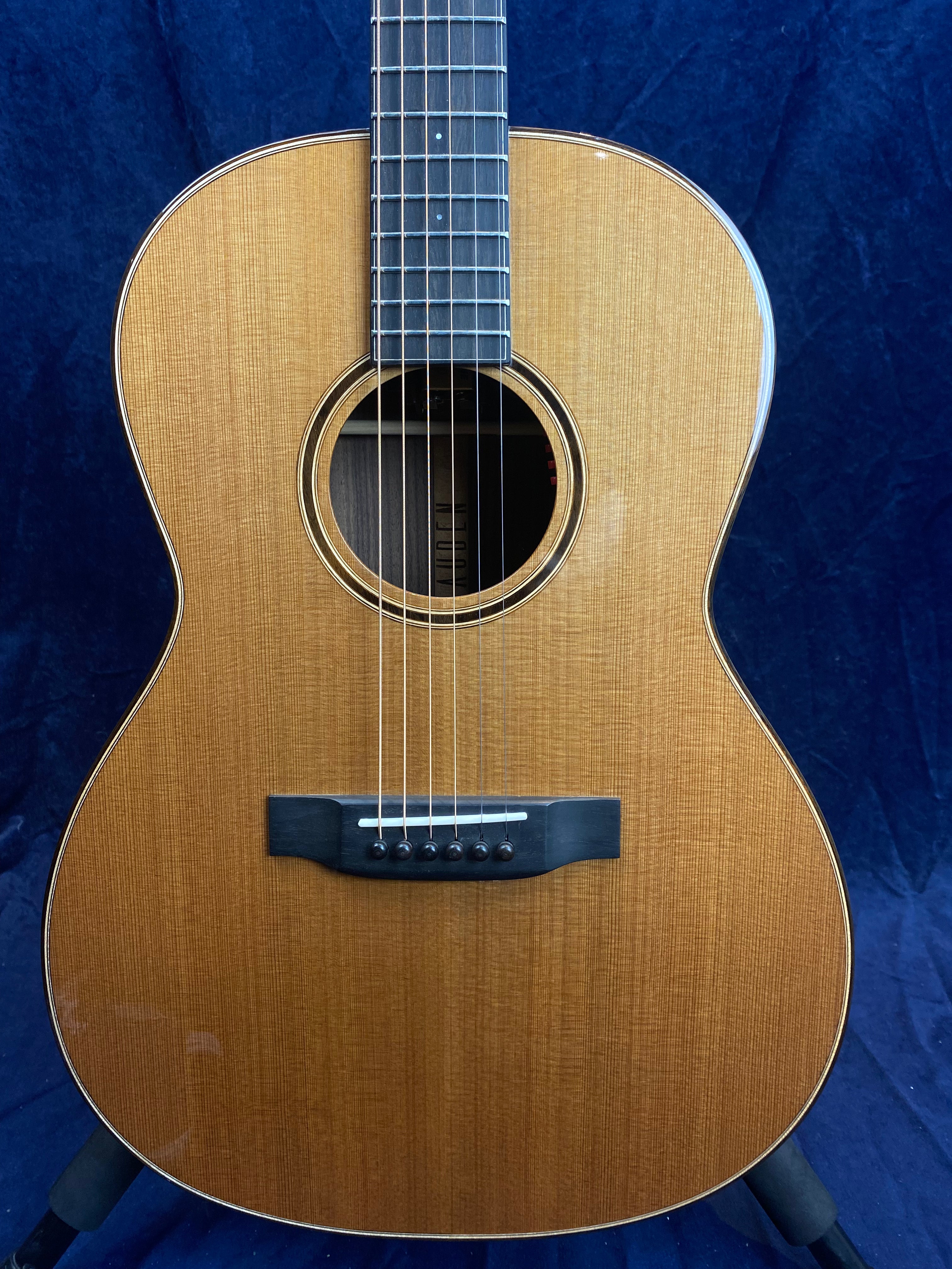 Light brown acoustic guitar with a dark wooden bridge, six strings, a round sound hole, and white edge binding. Blue background.
