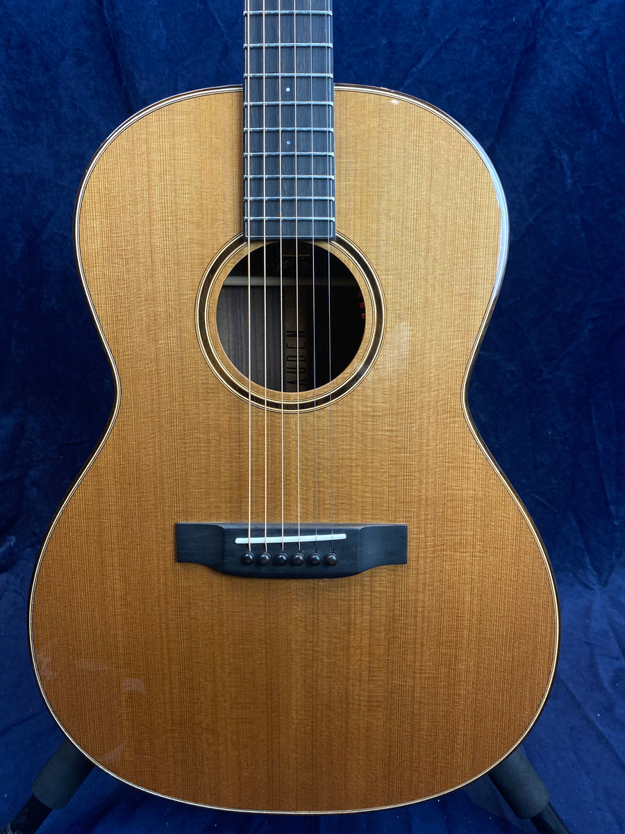 Light brown acoustic guitar with a dark wooden bridge, six strings, a round sound hole, and white edge binding. Blue background.
