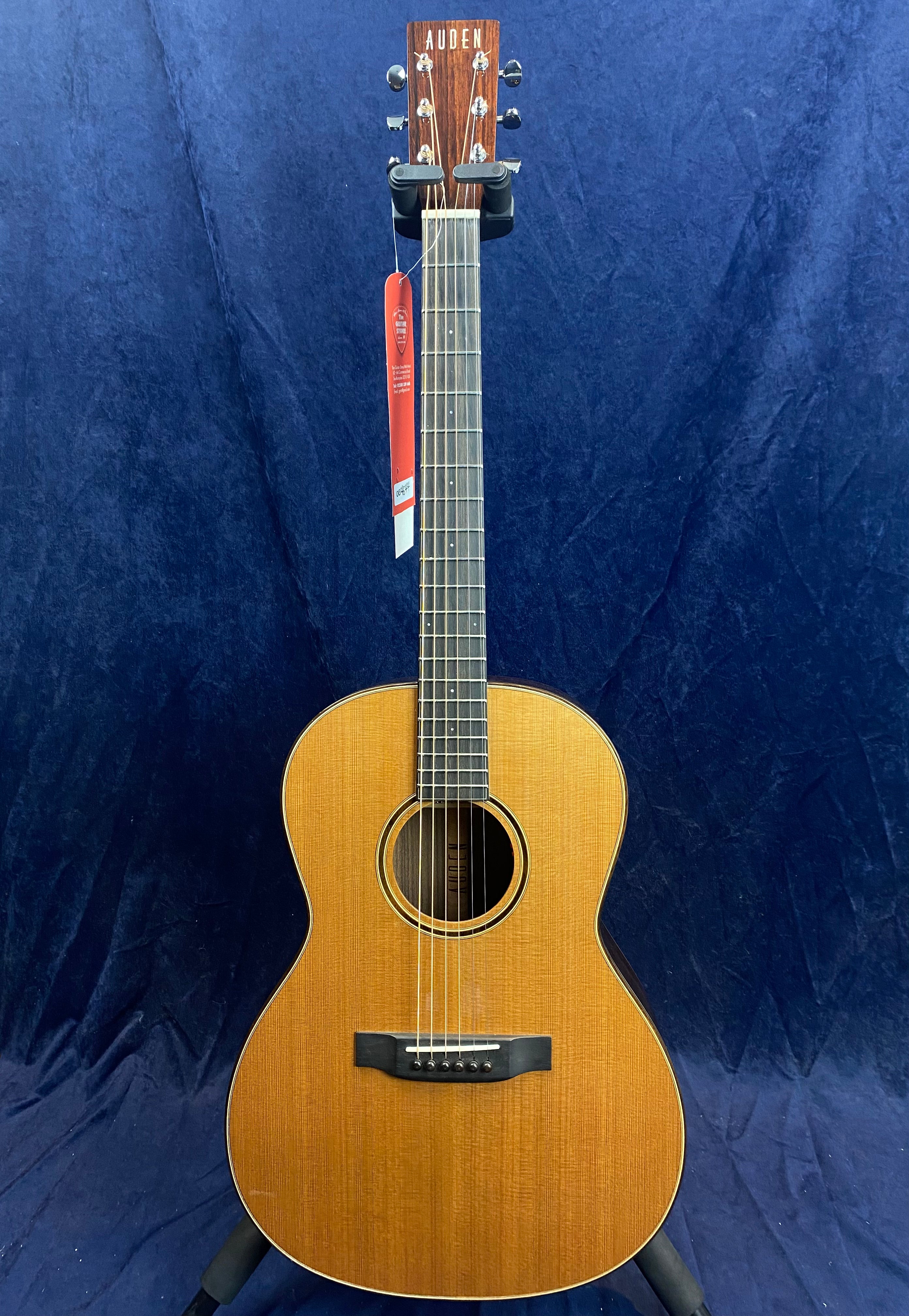Auden acoustic guitar with a natural wood finish, six strings, dark fretboard, and a circular sound hole, displayed upright against a dark blue background.