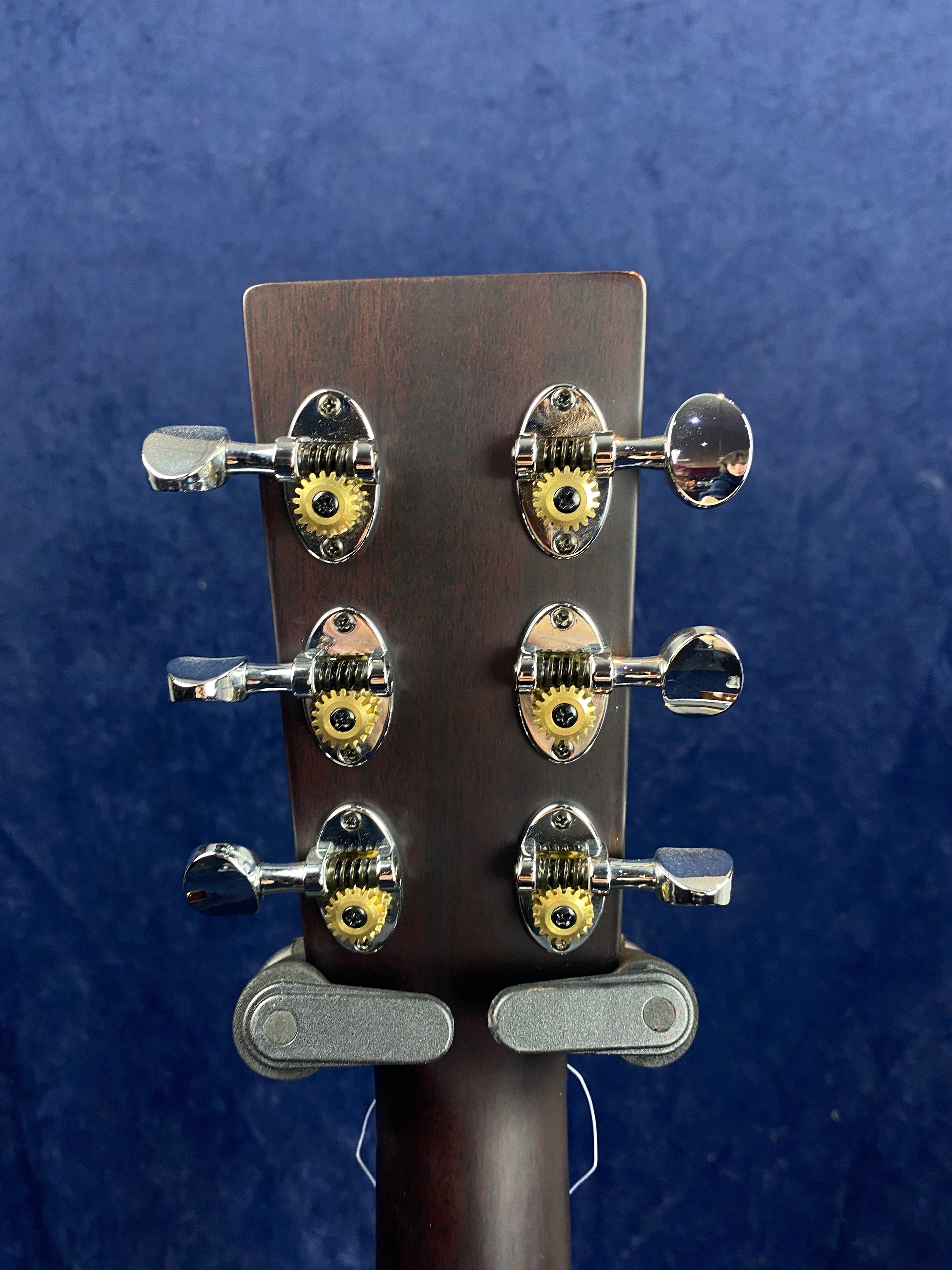 Six metal tuning pegs with exposed gears are mounted on the dark wooden headstock of an acoustic guitar against a deep blue background.