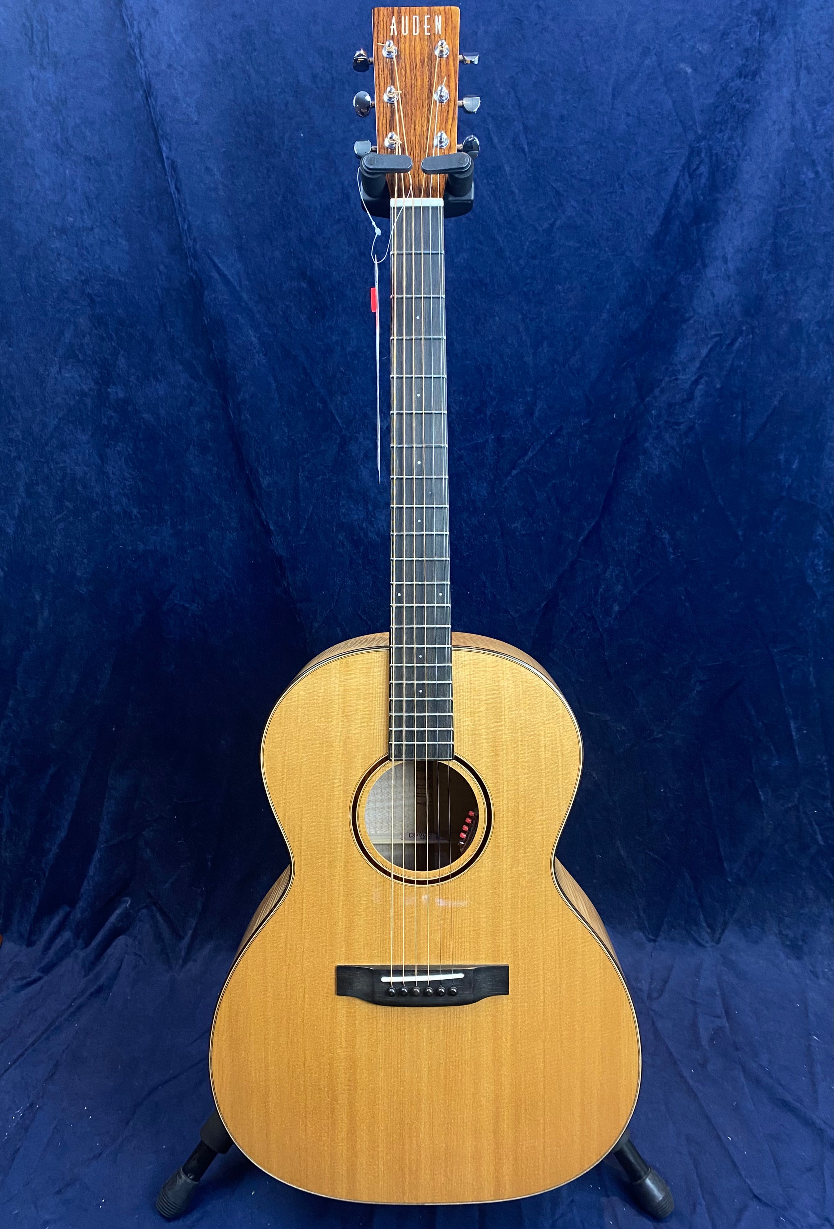 Light wood Auden acoustic guitar with six strings, dark fretboard, round soundhole, and black bridge, standing on a dark blue backdrop.