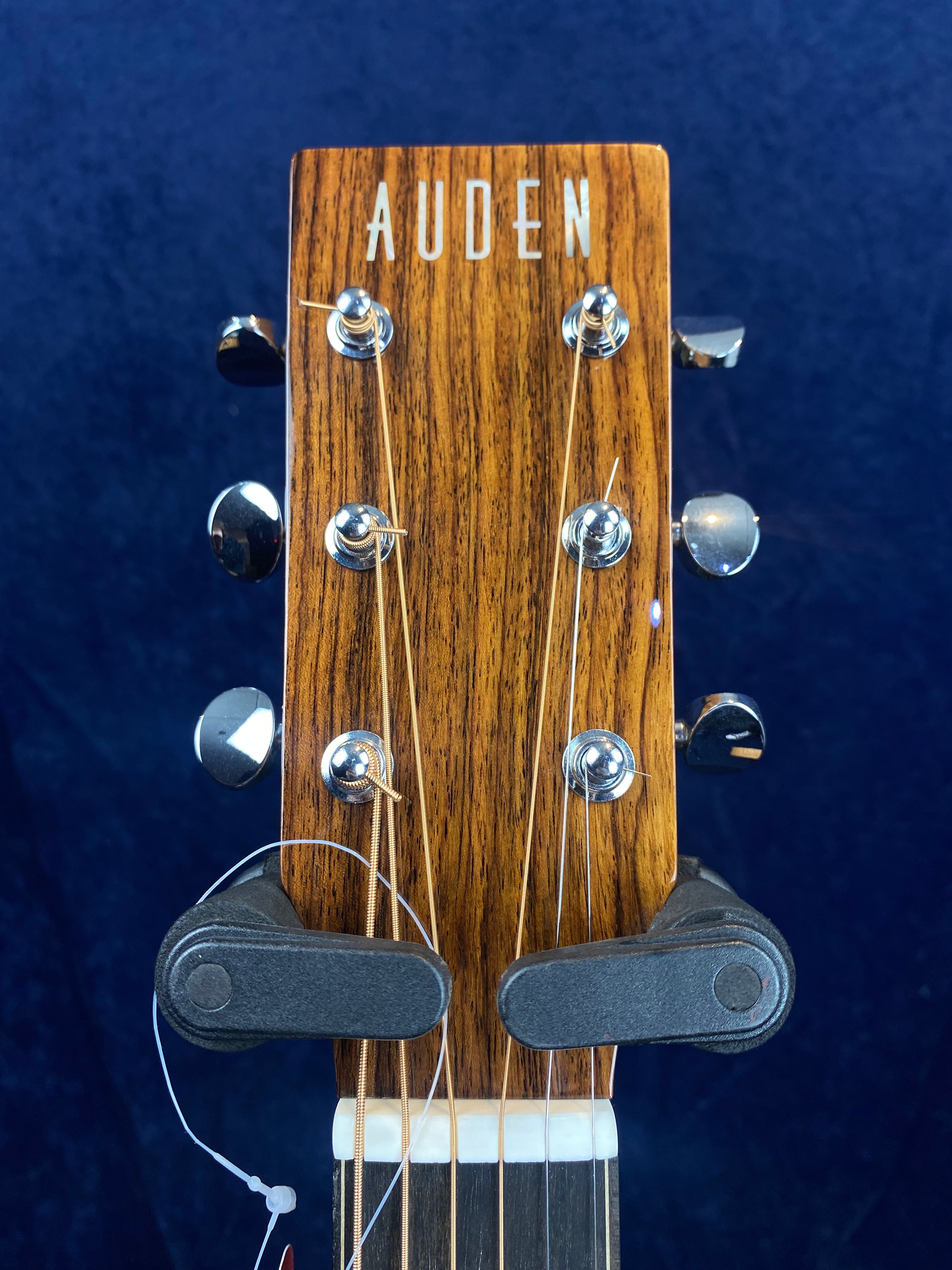 Auden acoustic guitar headstock with six metal tuning pegs, visible strings, and a dark wood finish against a blue background.